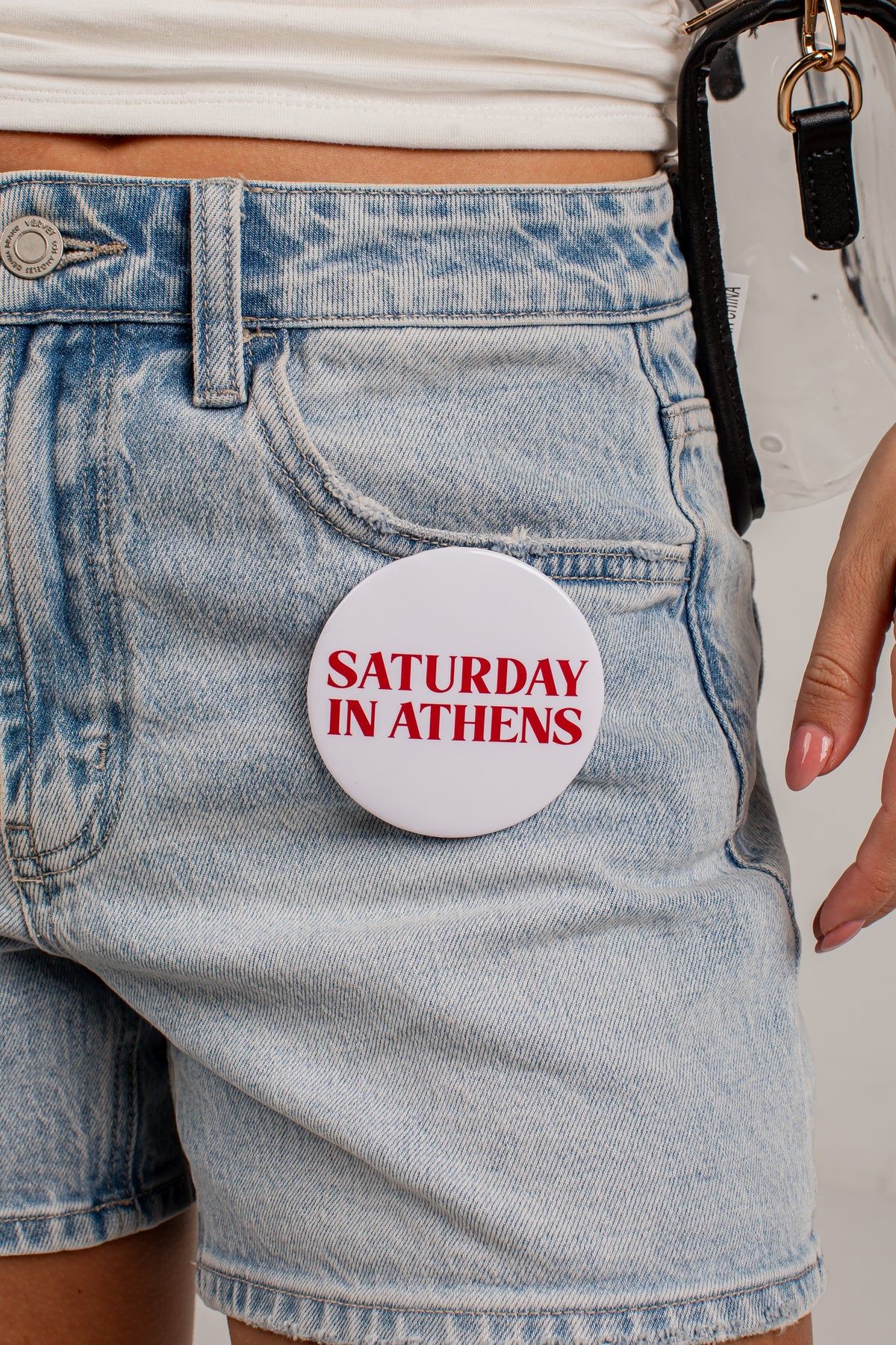 A close-up of a person wearing light-wash denim shorts and a white top, with a round white pin or badge attached to the pocket. The badge has red text that reads “SATURDAY IN ATHENS.