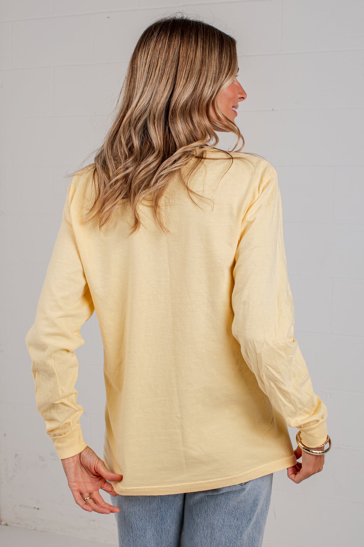 A woman with blonde hair is shown from the back wearing a pale yellow long-sleeve top and light blue jeans against a white wall.