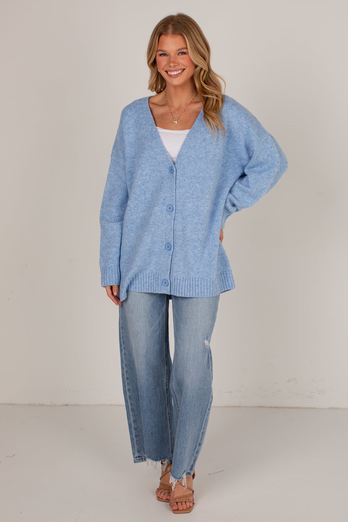 A woman modeling a light blue knit cardigan sweater over a white top and wide-leg light blue jeans, standing against a plain off-white studio background.