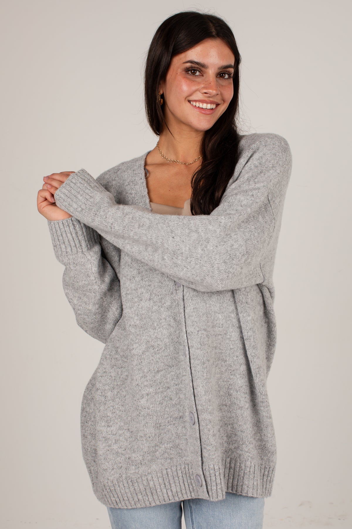 A woman modeling an oversized light gray knit cardigan sweater with a V-neck and ribbed cuffs and hem, paired with light blue jeans against a plain light gray studio background.