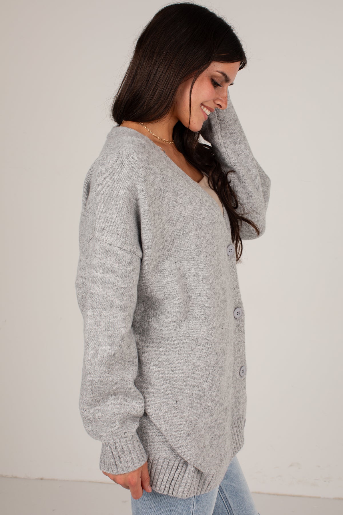 A woman wearing a light gray heathered knit cardigan sweater with a V-neck and button-front closure, paired with light blue jeans, photographed against a plain off-white wall.