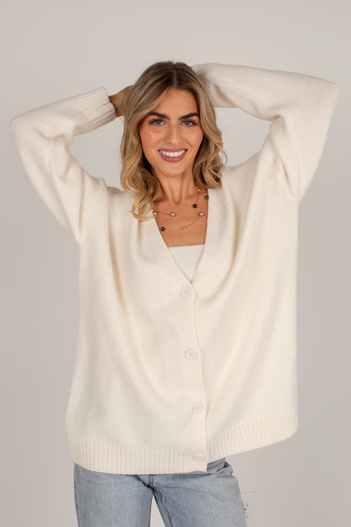 A woman wearing an oversized cream-colored knit cardigan sweater with buttons, paired with light blue jeans, posing with her hands behind her head against a solid light gray background.