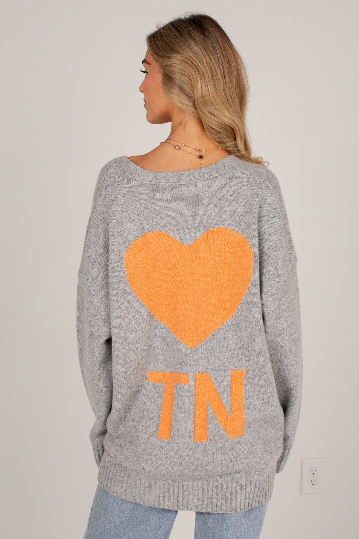 A woman shown from the back wearing a heather gray knit sweater with a large orange heart and the letters &