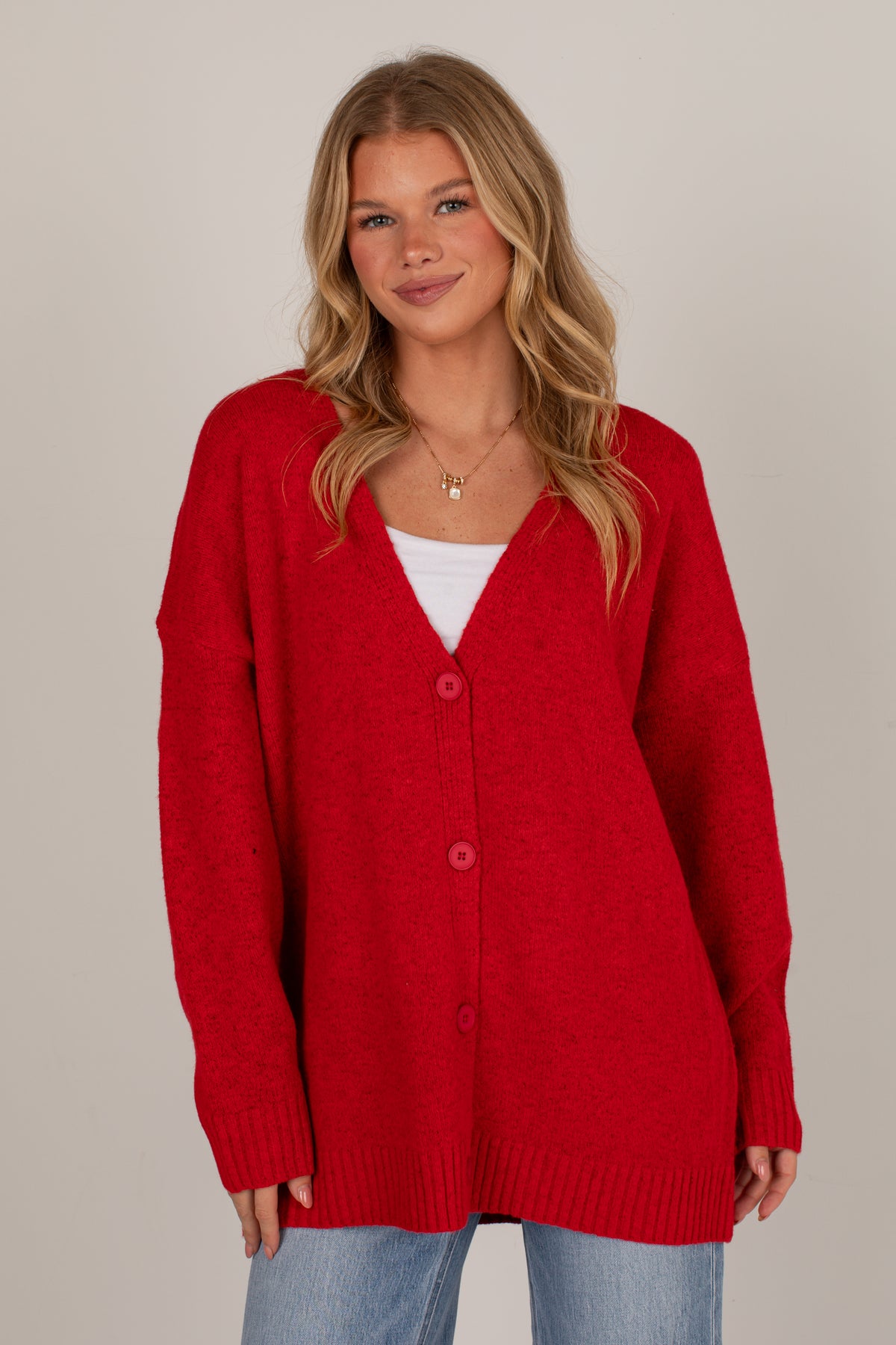 A woman wearing a bright red knit cardigan over a white top and light blue jeans, standing against a plain light gray studio background.