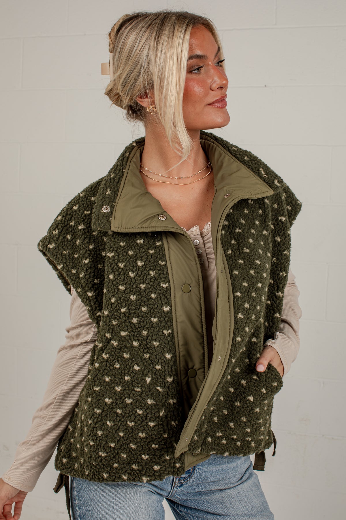 A woman wearing an olive green fleece vest with a beige and green pattern, layered over a beige long-sleeve top and blue jeans, standing against a white brick wall.