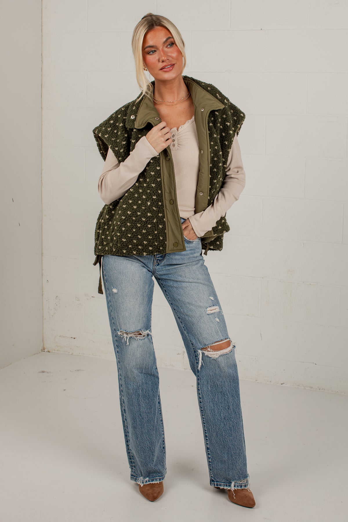 A woman modeling a sleeveless olive green vest with white polka dots over a beige top, paired with distressed blue jeans and brown boots, standing against a white brick wall.