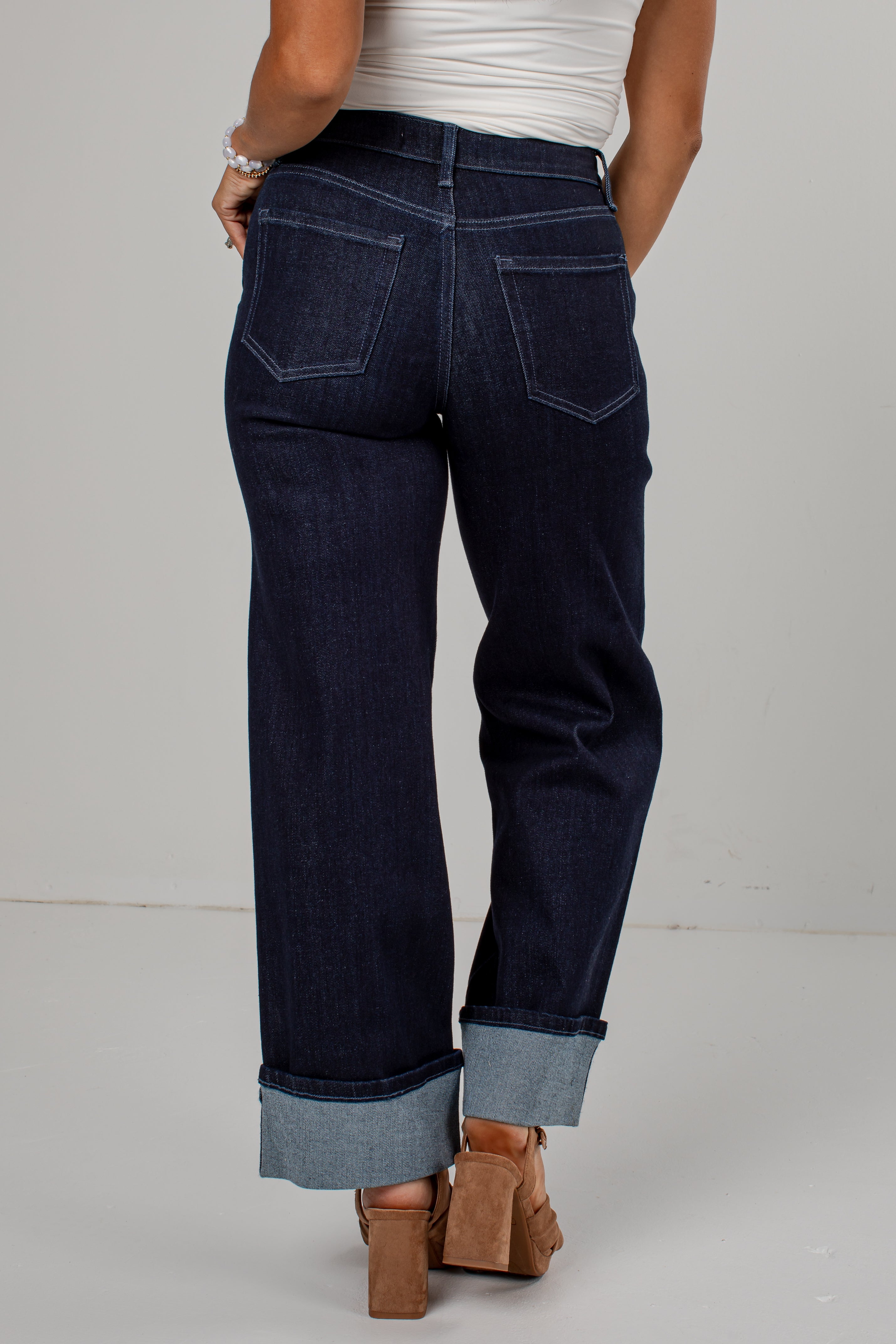 Rear view of a person wearing dark indigo denim jeans with rolled cuffs, paired with a white top and brown block-heel sandals, against a light gray studio background.