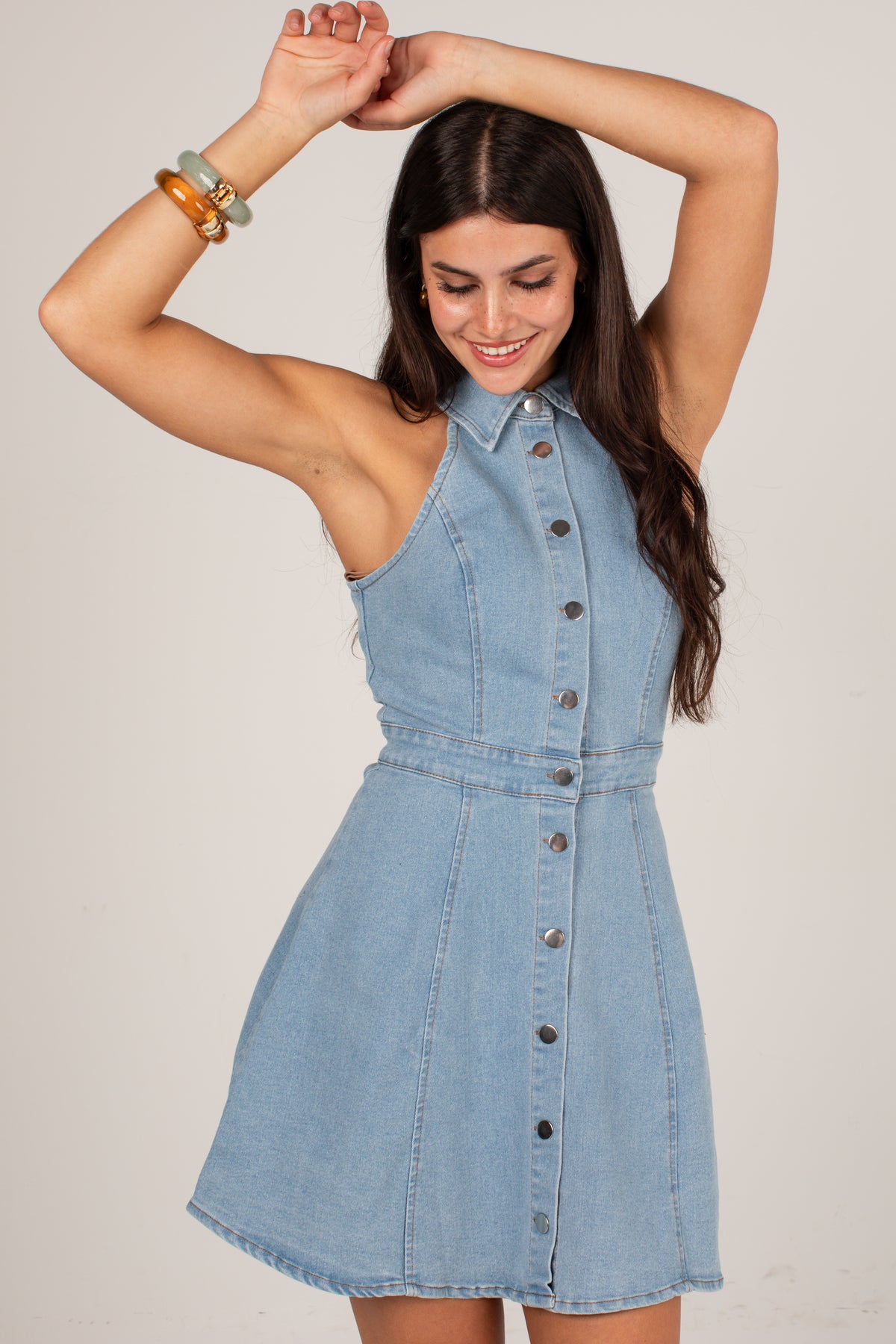 A woman modeling a light blue denim halter dress with a button-down front and a flared skirt, posed against a plain light beige studio background.