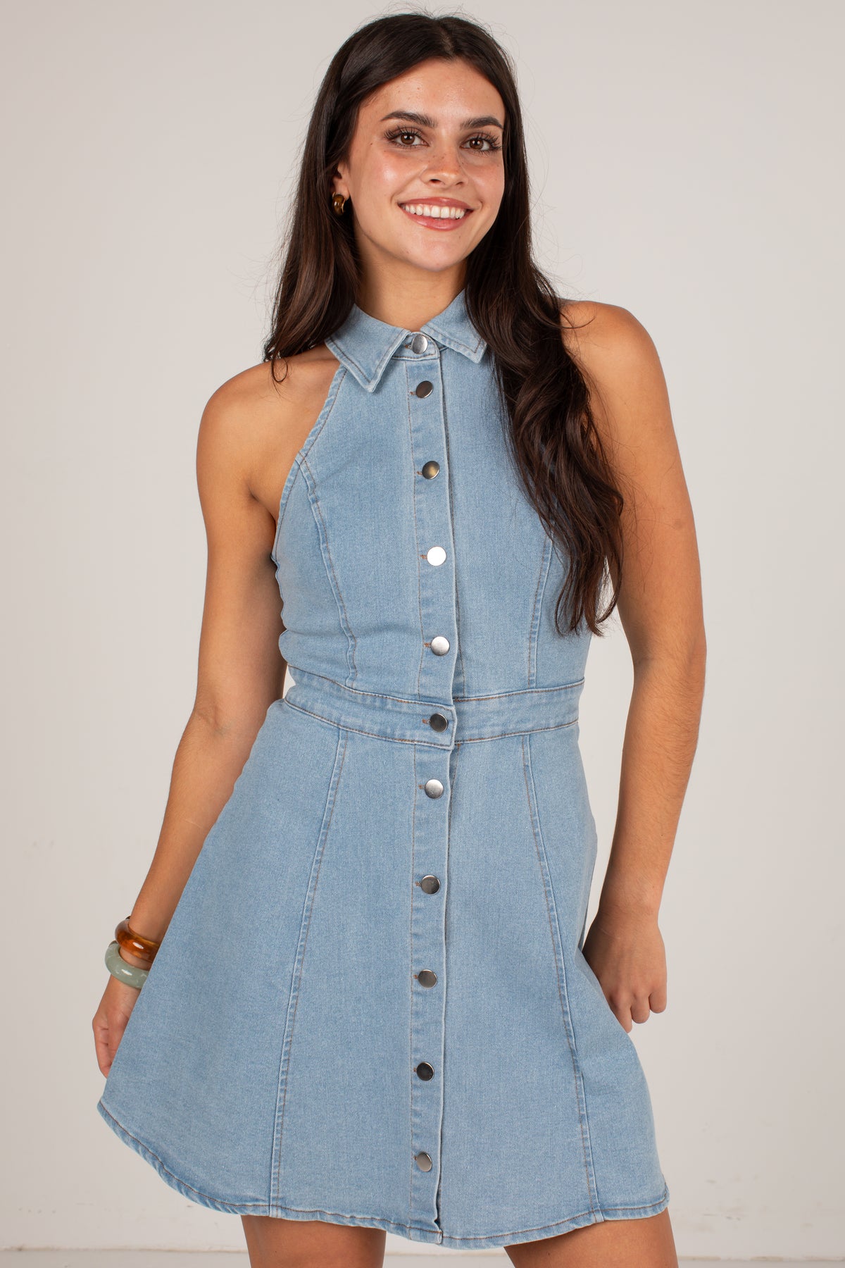 A woman modeling a sleeveless light blue denim halter dress with a button-down front and a flared skirt, set against a plain light gray studio background.