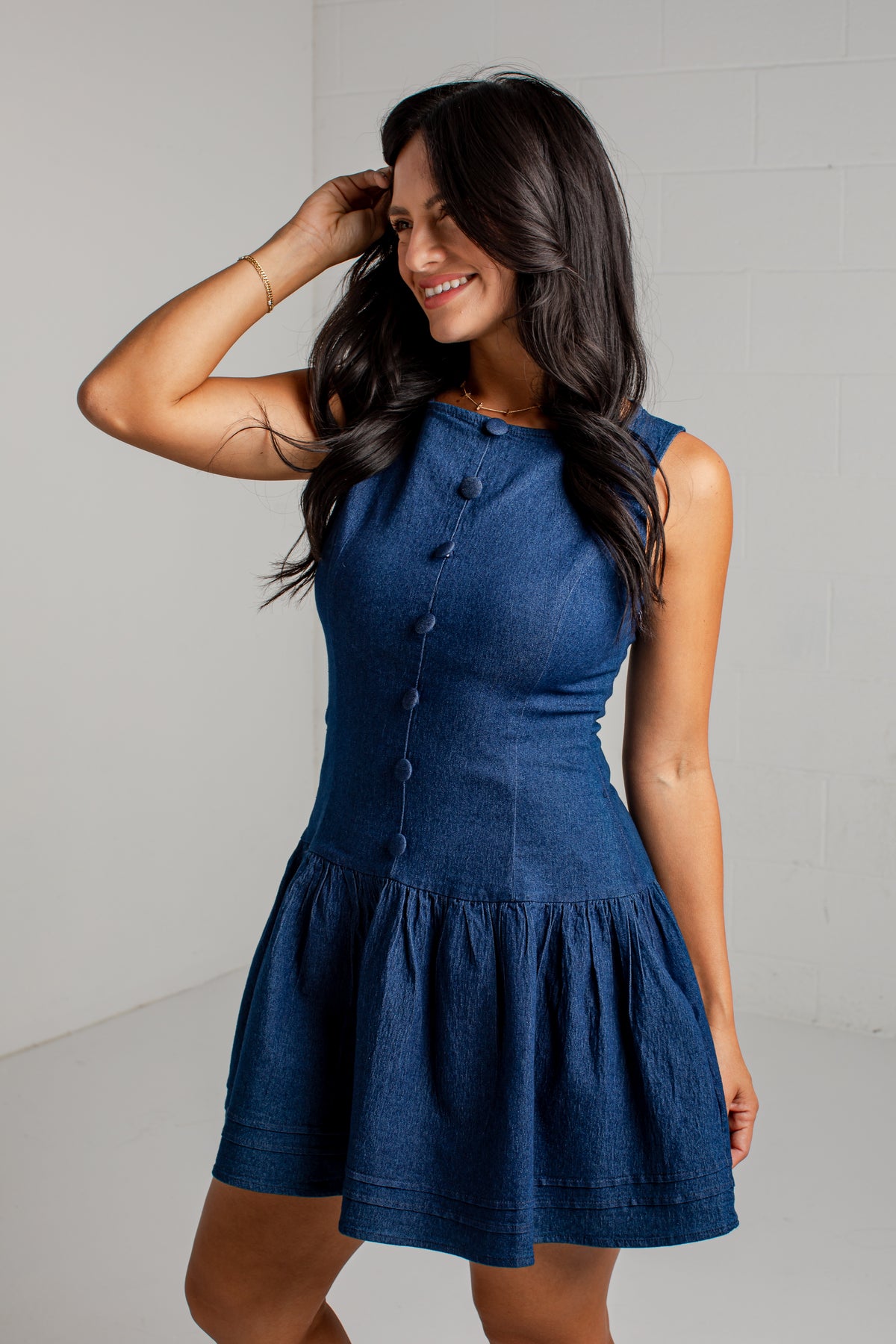 A woman modeling a sleeveless blue denim dress with a buttoned neckline and a flared skirt, standing against a light gray wall.