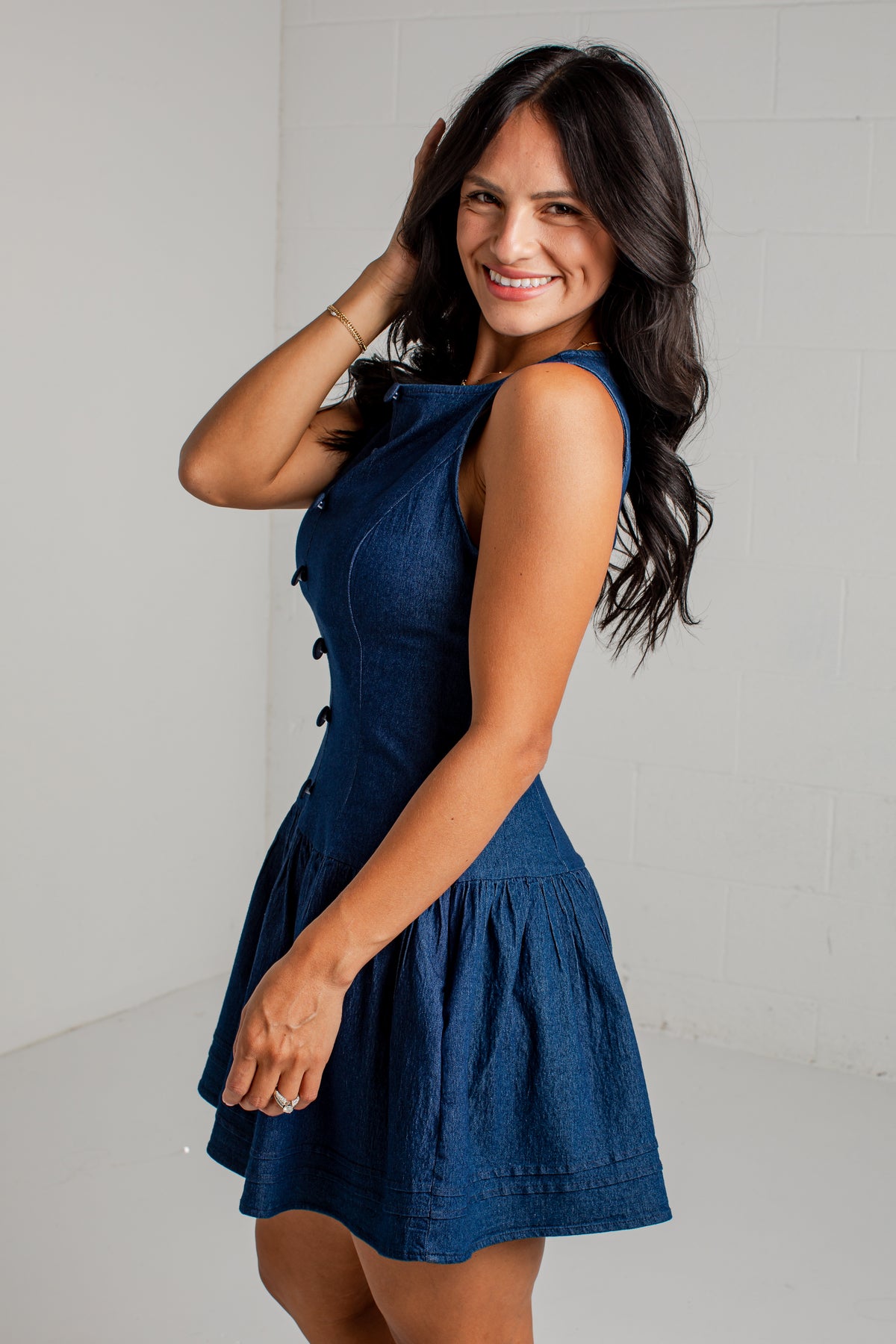 A woman modeling a sleeveless dark blue mini dress with a flared skirt and decorative buttons down the front, posed against a light gray brick wall.