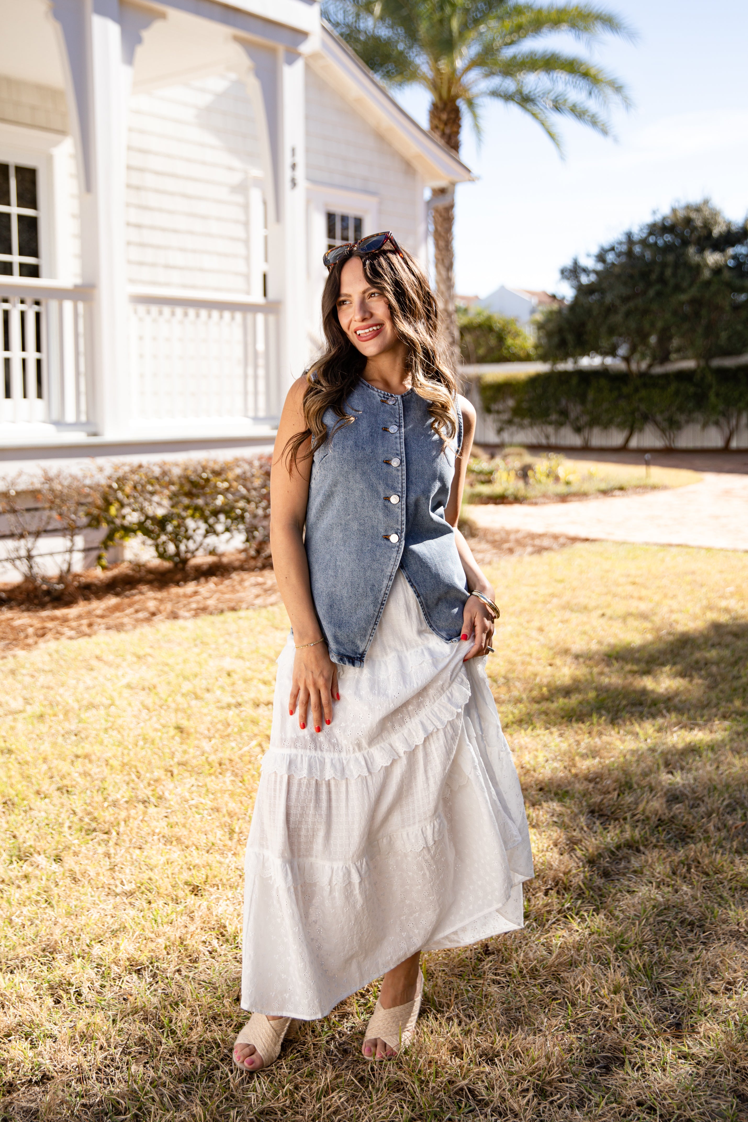 Woman in a white dress and denim vest standing in a grassy area with a white building and palm tree in the background.