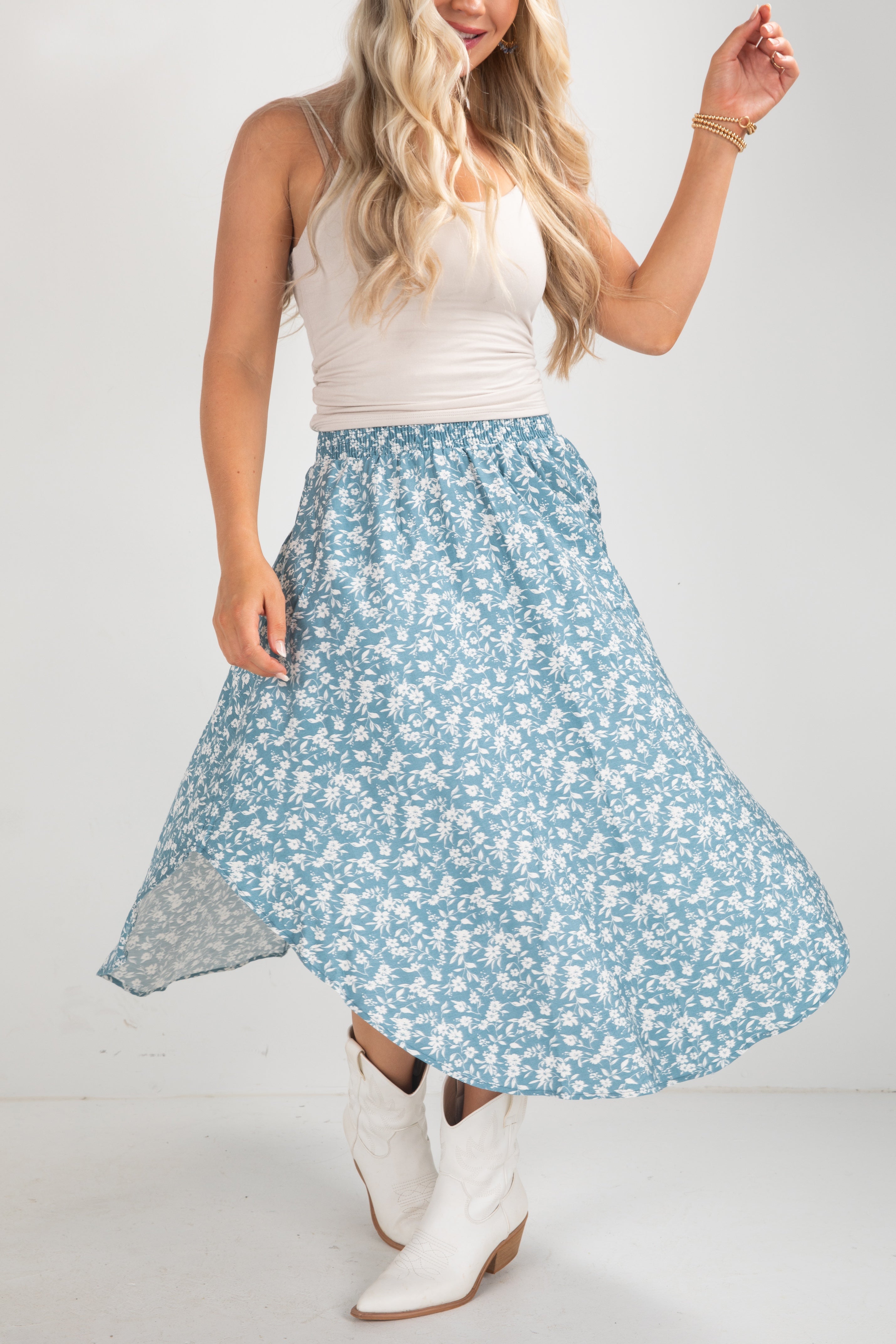 Woman wearing a white tank top and blue floral skirt with white boots on a plain background
