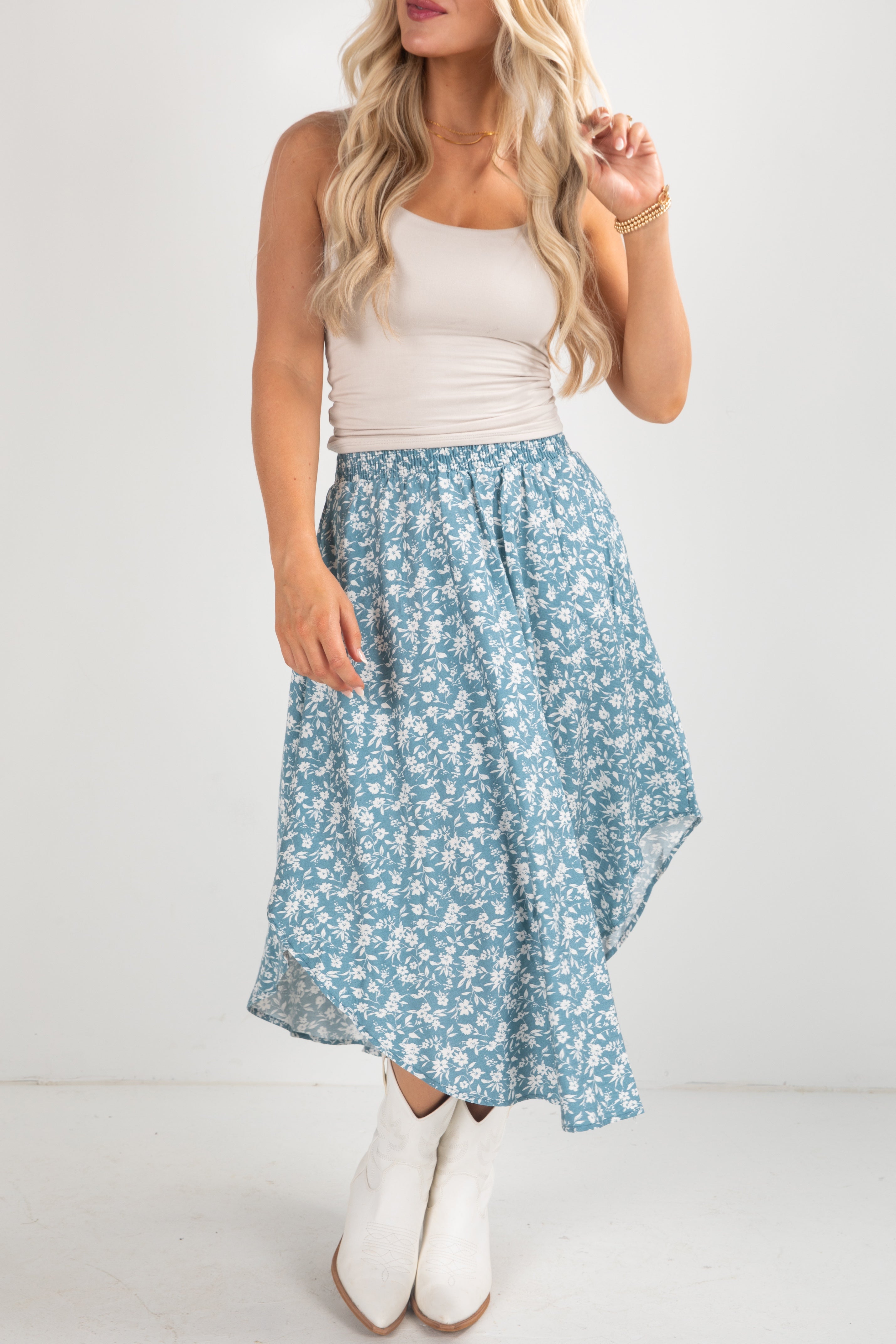 Woman wearing a white tank top and blue floral skirt with white boots on a light background