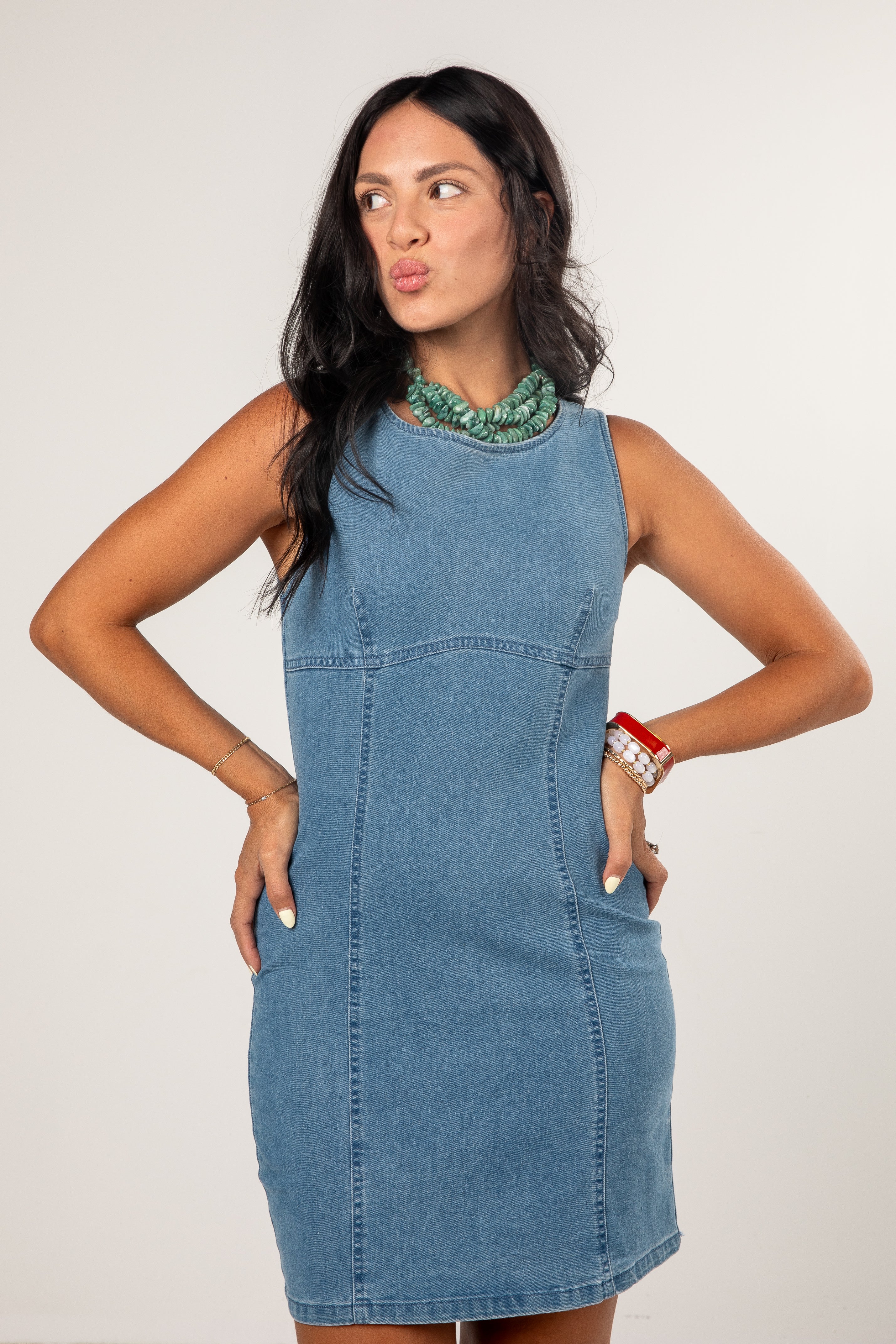 A woman modeling a sleeveless light blue denim dress with side pockets and a green beaded collar, posing against a light gray studio background.
