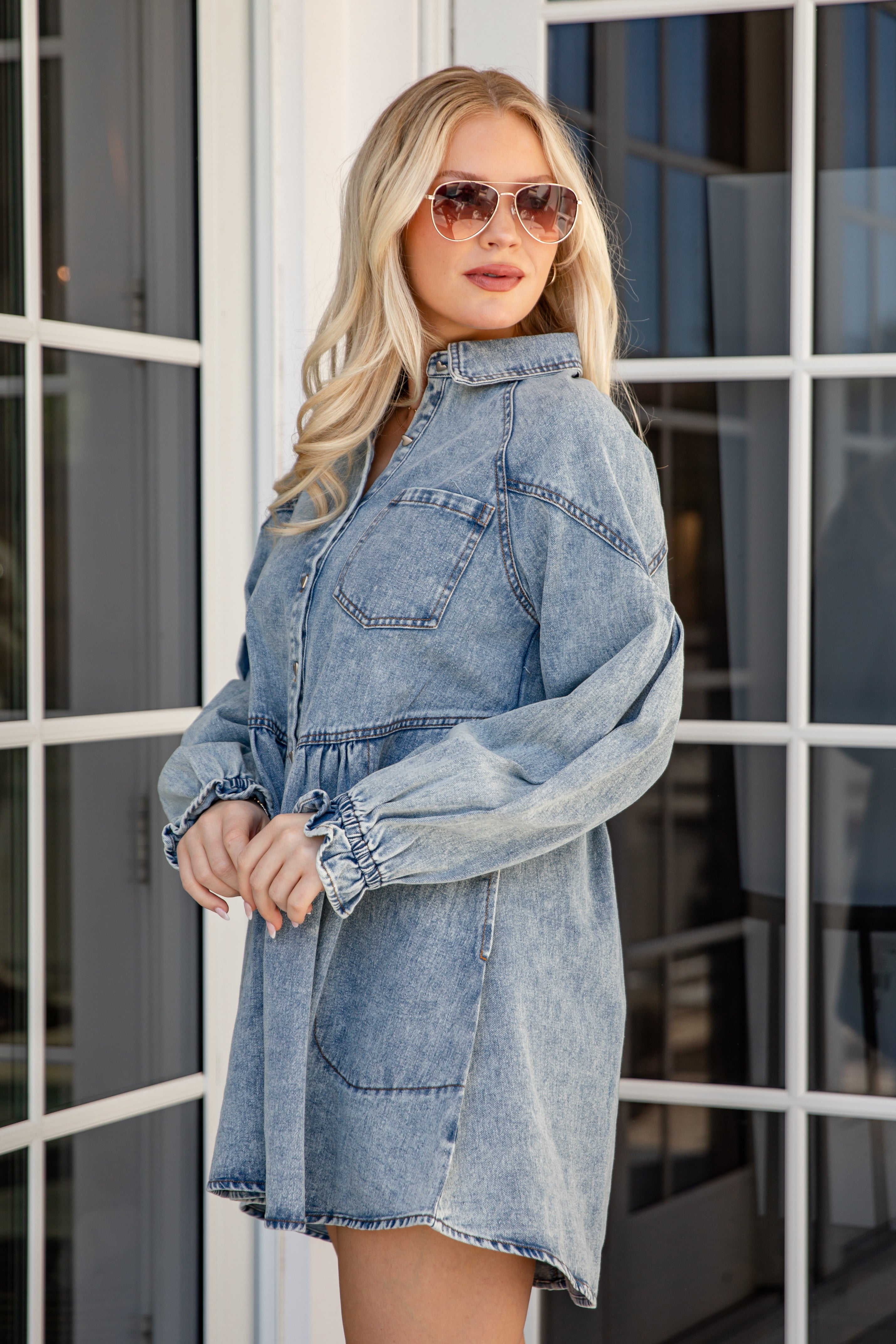 Woman wearing a denim dress and sunglasses in front of a glass door.