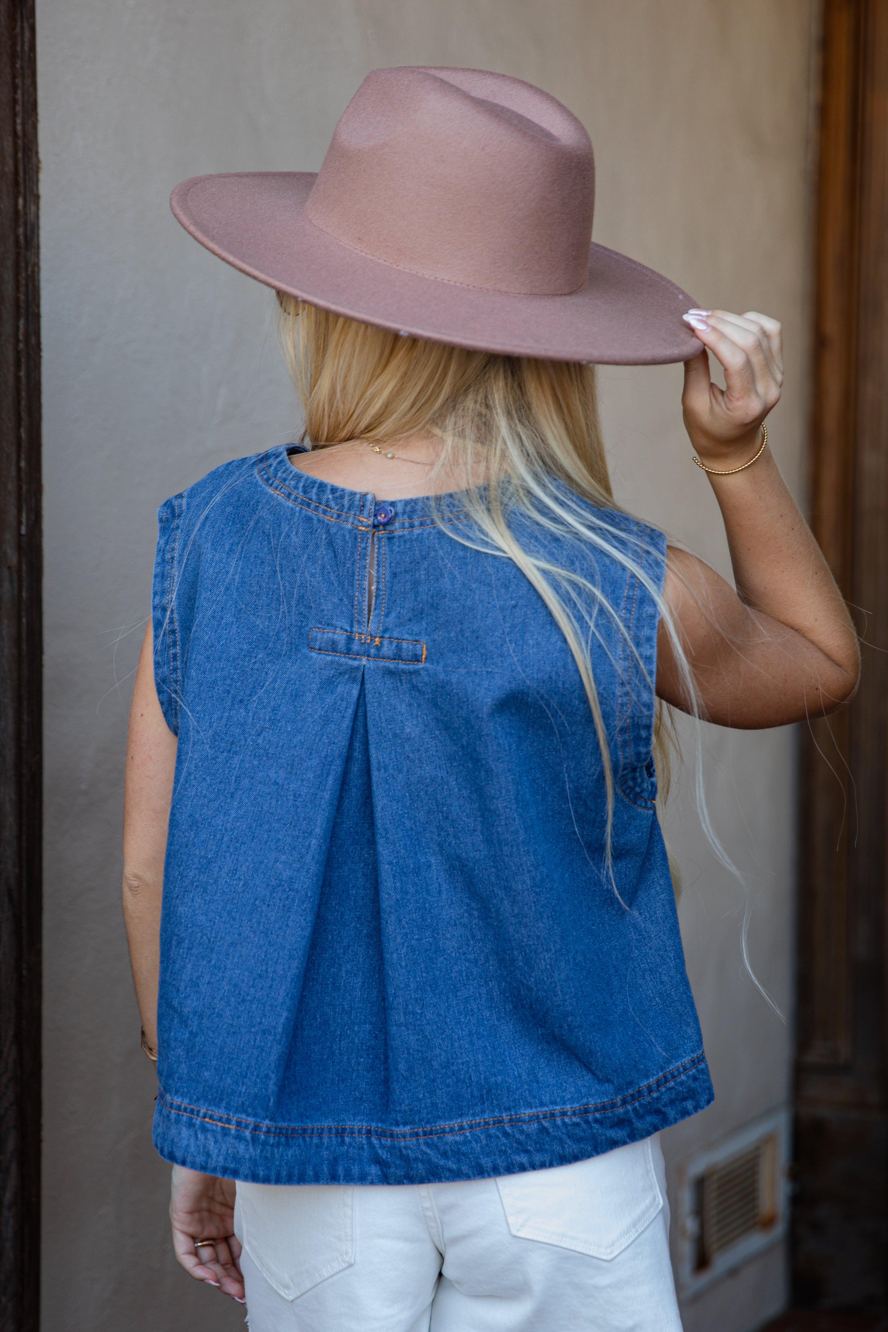 A woman viewed from behind wearing a sleeveless blue denim top with a gathered back seam, white pants, and a dusty pink wide-brim hat, standing against a neutral beige wall.
