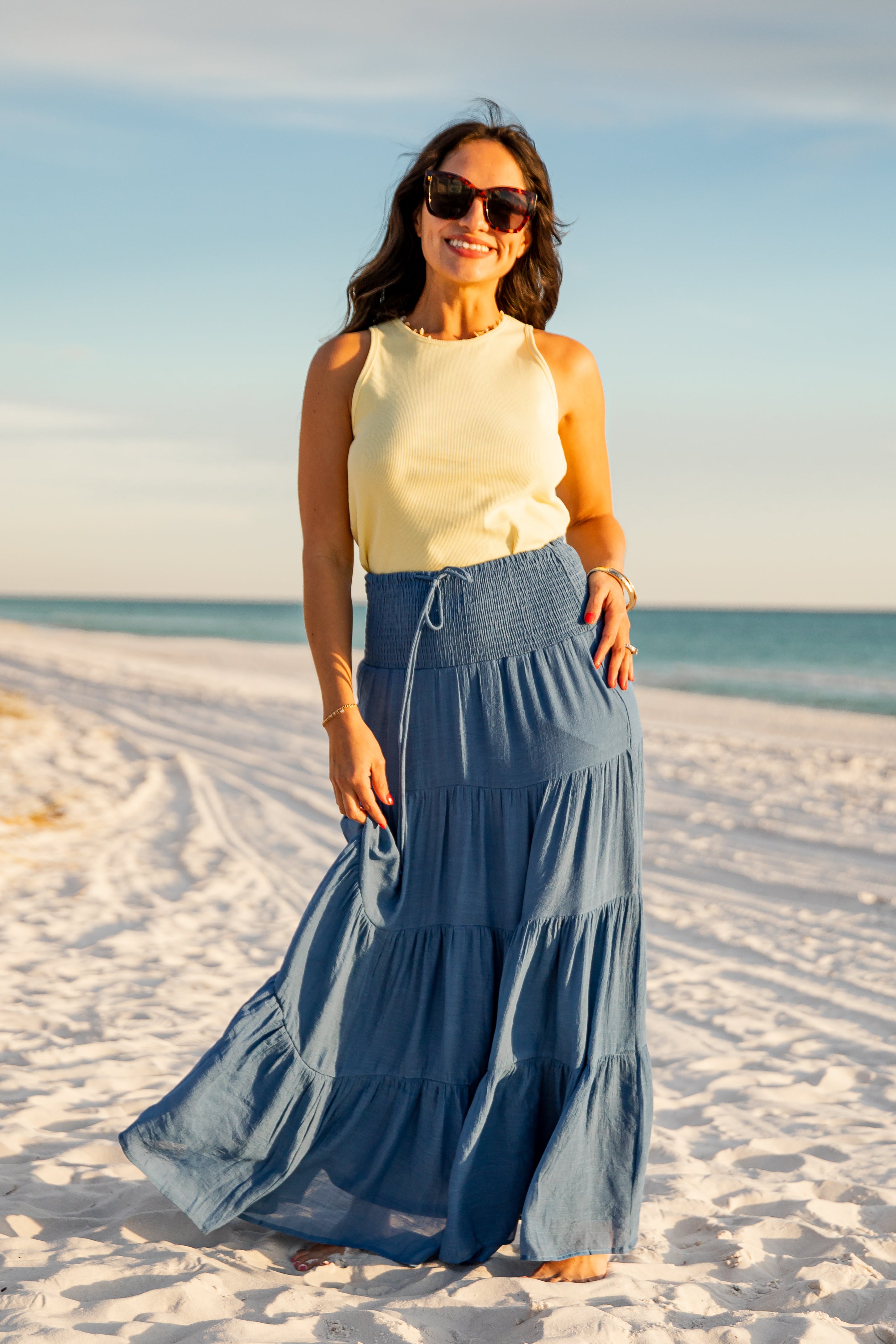 Woman in a yellow top and blue skirt standing on a sandy beach with ocean view.