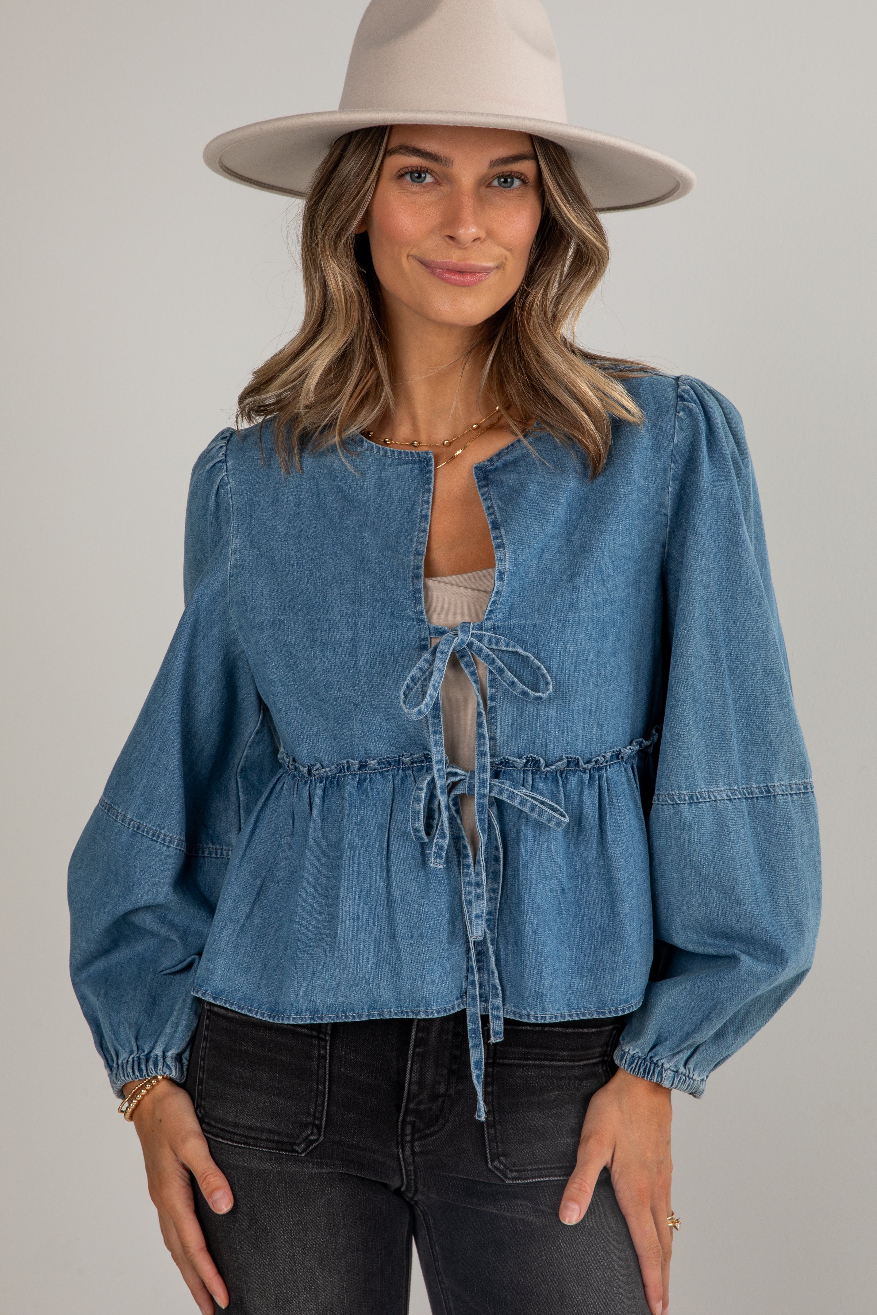 Woman wearing a blue denim blouse with puffed sleeves and a beige hat against a gray background