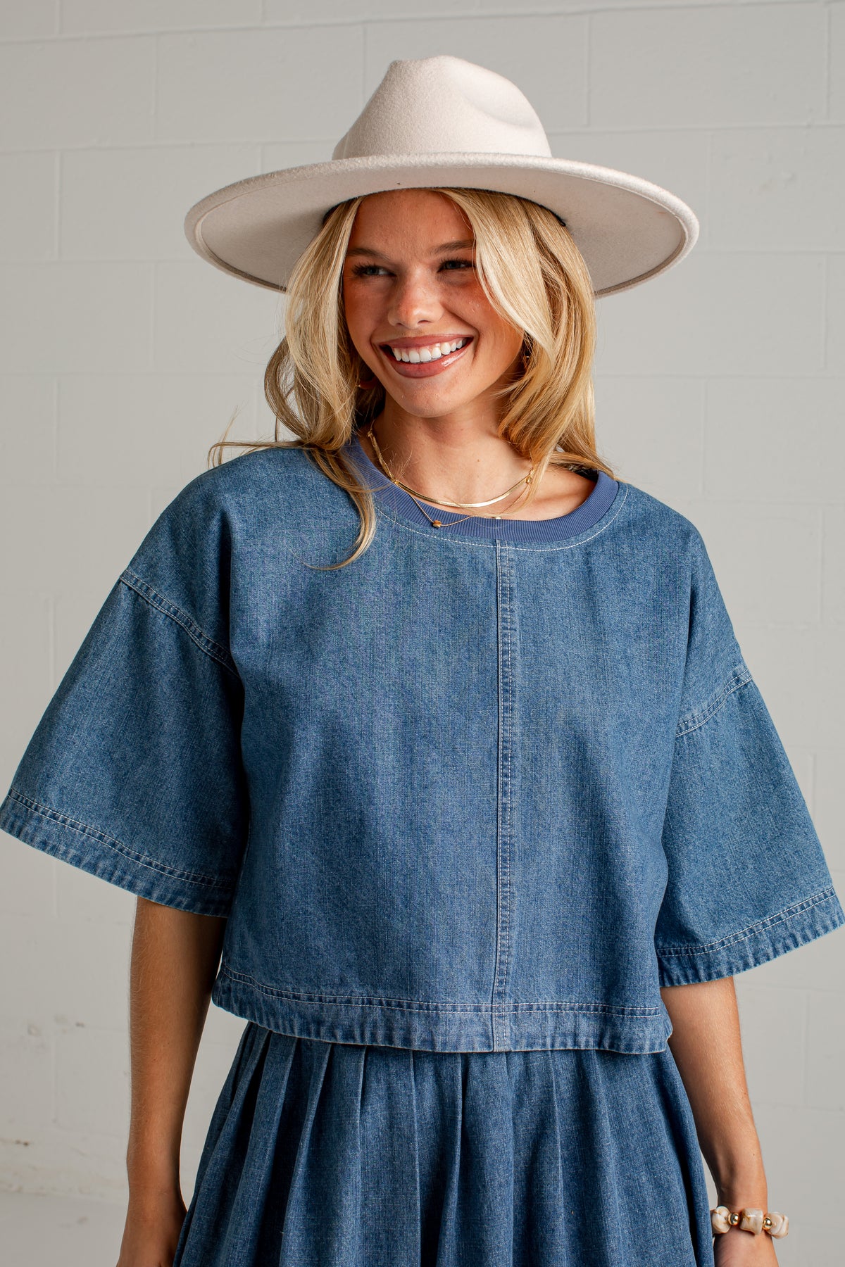 A smiling blonde woman wearing a light beige wide-brim hat and a blue denim short-sleeve top with a matching pleated skirt, standing against a light gray brick wall.