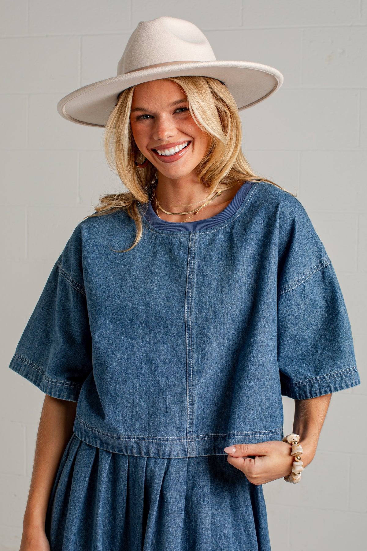 A smiling blonde woman wearing a blue denim short-sleeve top and matching pleated skirt, paired with a wide-brimmed cream hat, against a light gray brick wall.