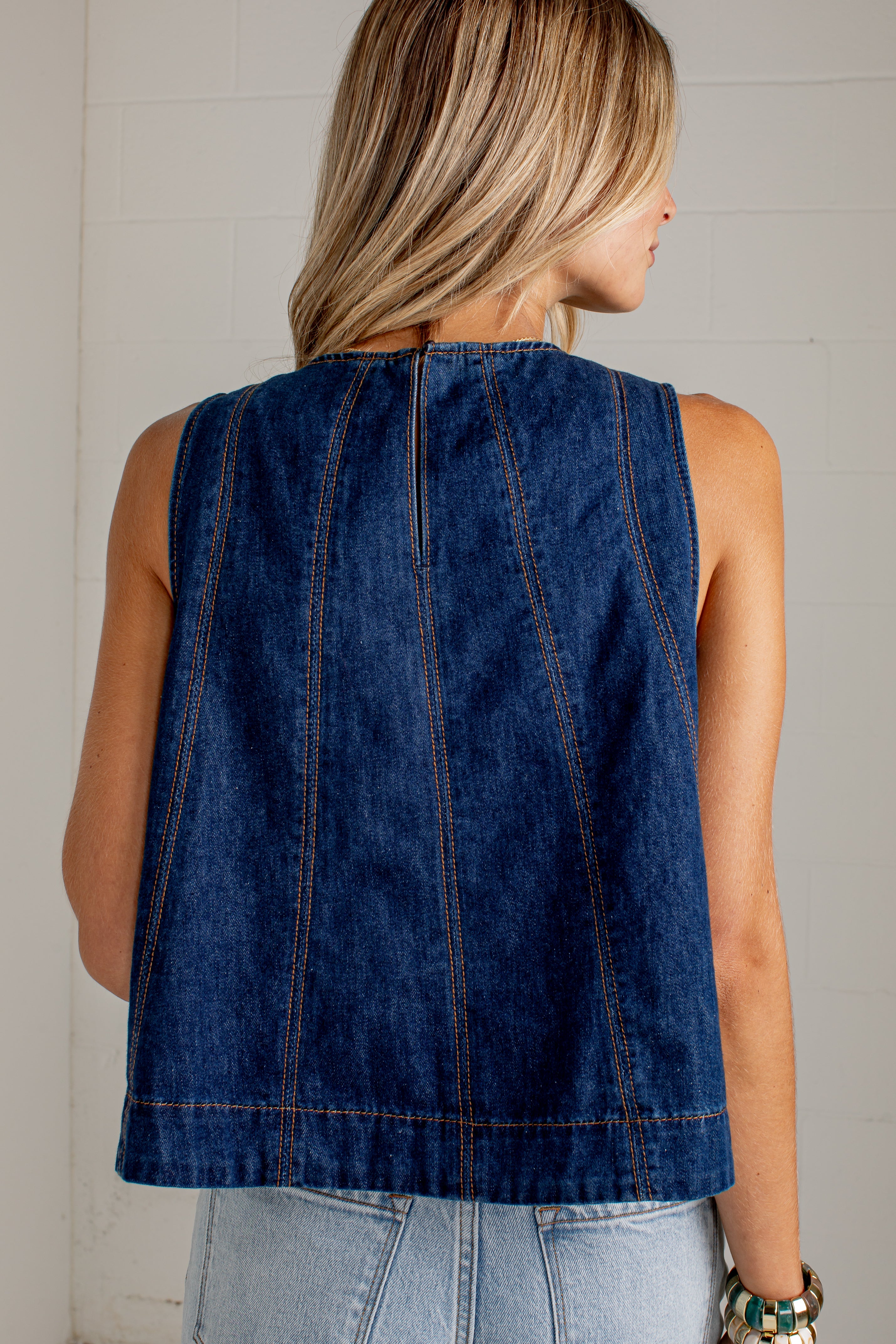 A woman shown from the back wearing a sleeveless dark blue denim vest with vertical seam detailing, paired with light blue jeans against a white brick wall.