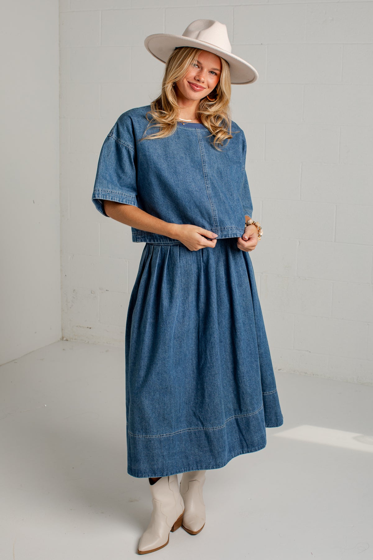 A woman modeling a blue denim midi dress with short sleeves and a gathered waist, styled with a beige wide-brim hat, beige boots, and gold jewelry against a white studio background.