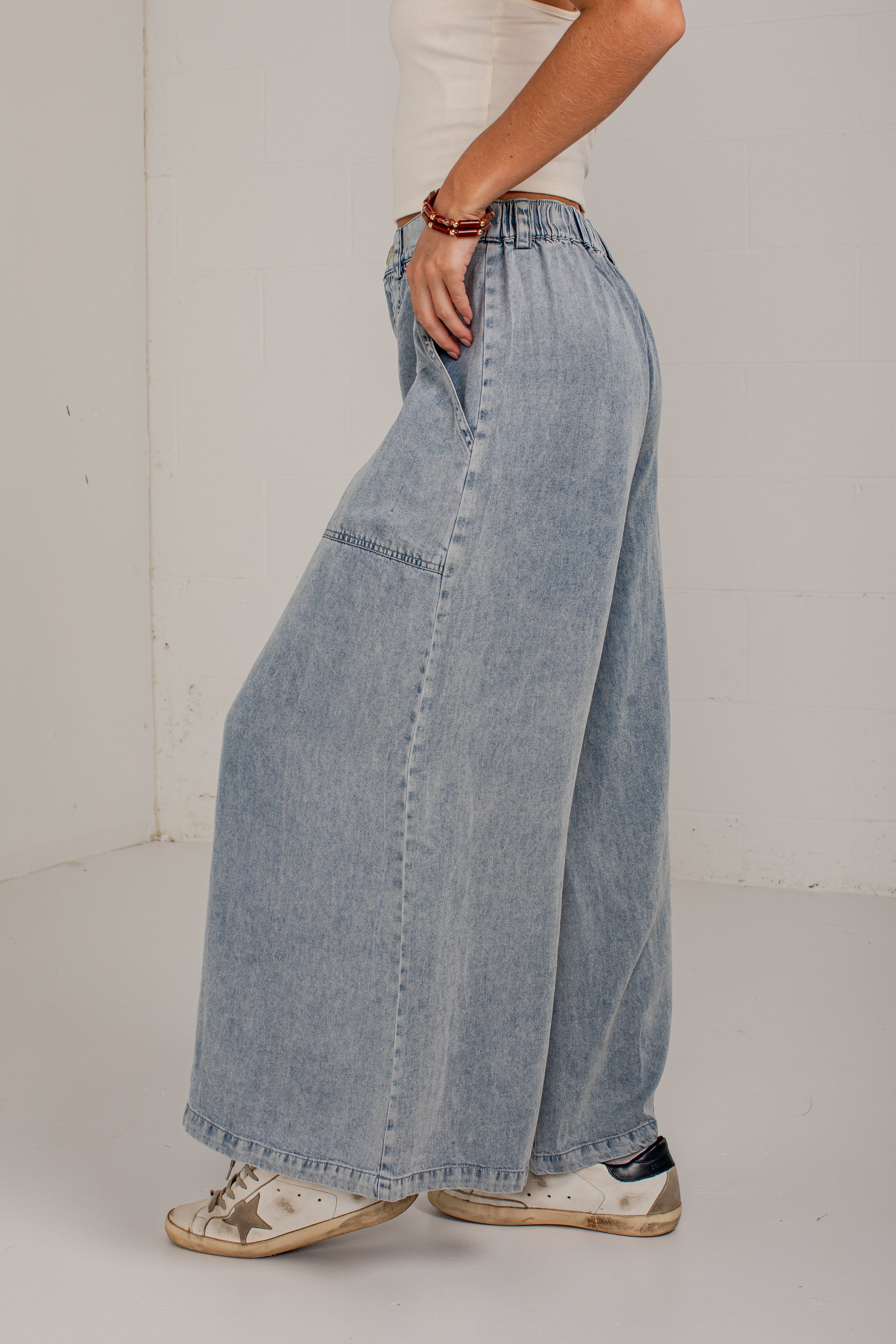 A person wearing light-wash wide-leg denim pants, shown from the waist down in a studio setting with a white wall and floor.