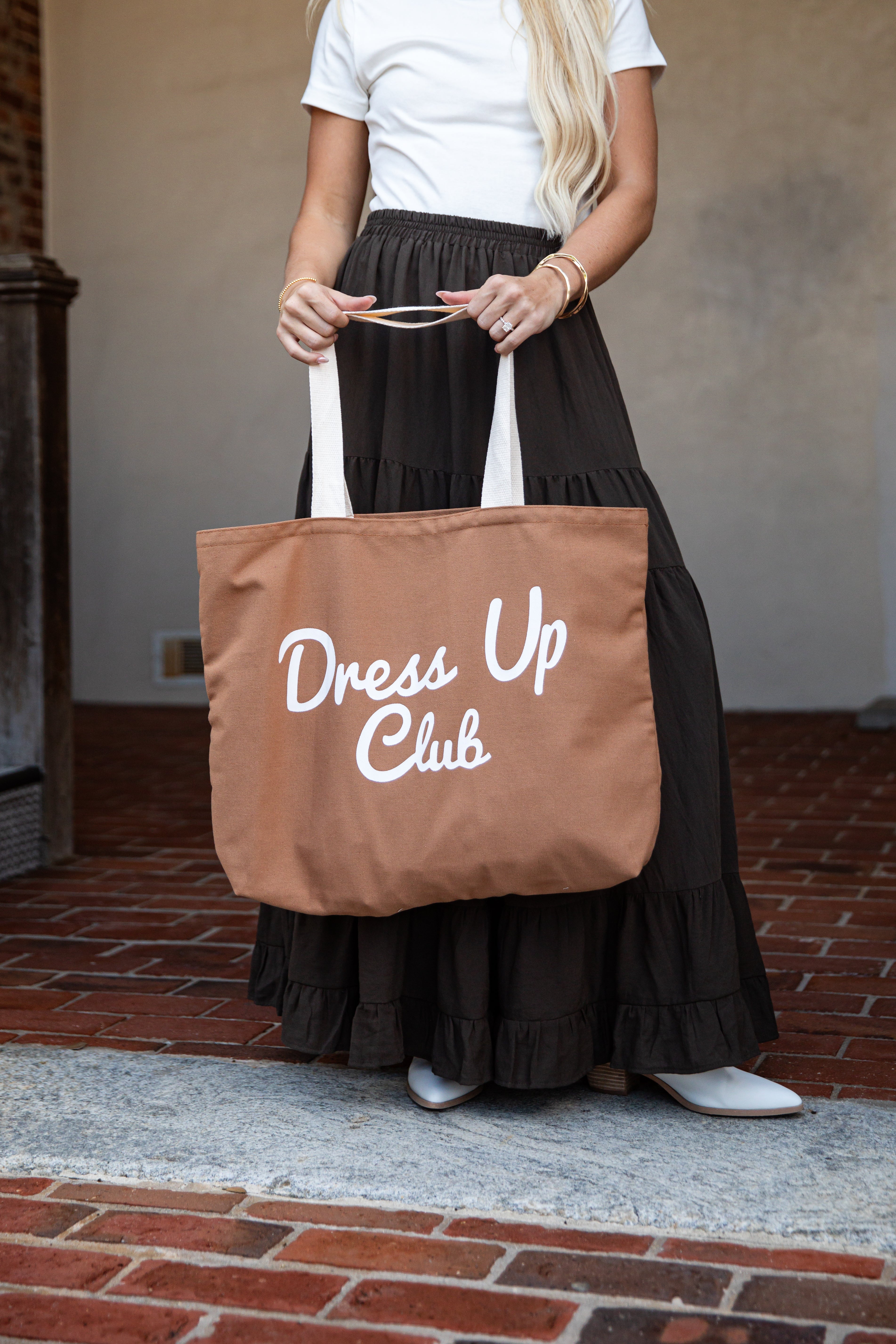 A woman wearing a white t-shirt and a long black ruffled skirt holds a brown canvas tote bag with white handles and the words &