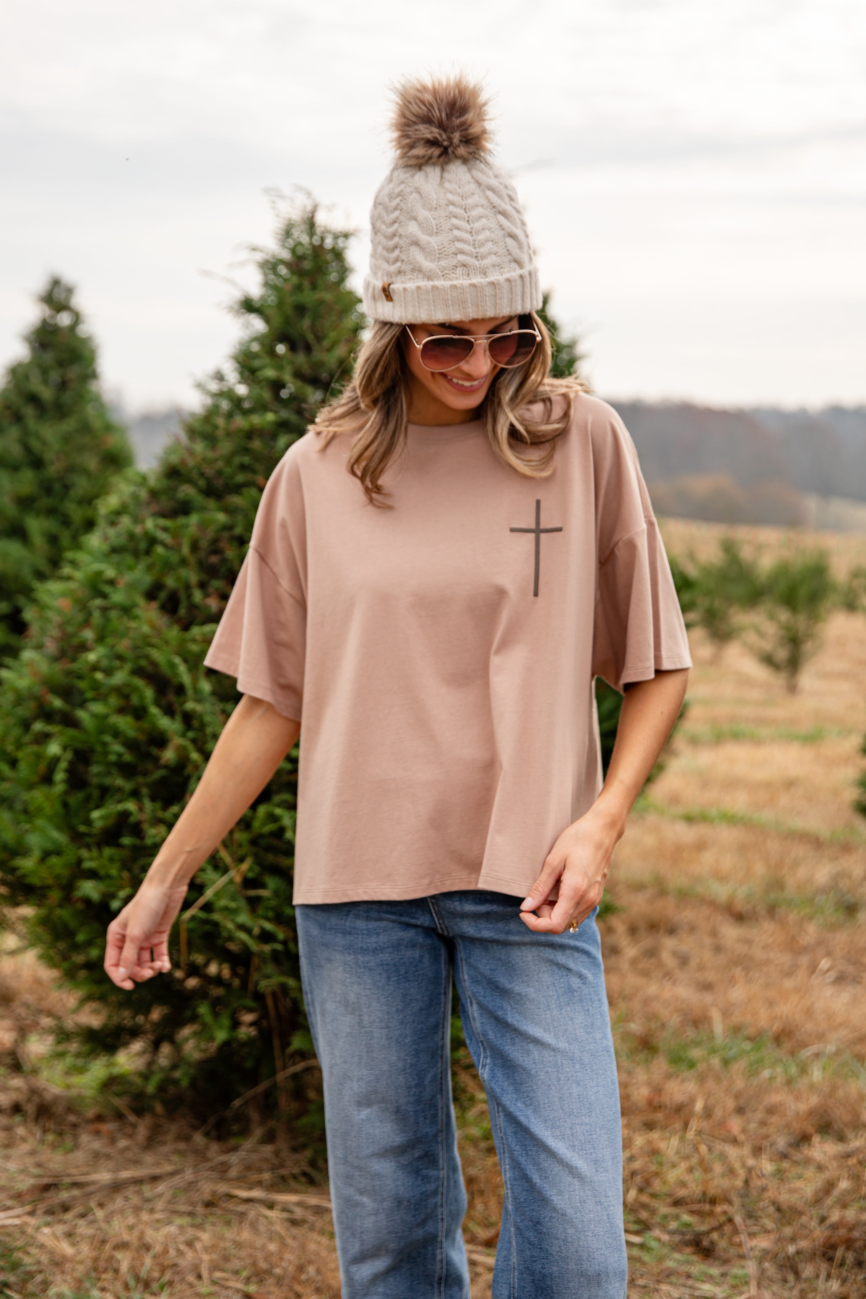 Woman wearing a beige oversized t-shirt with a cross design, blue jeans, and a knit hat in a field.