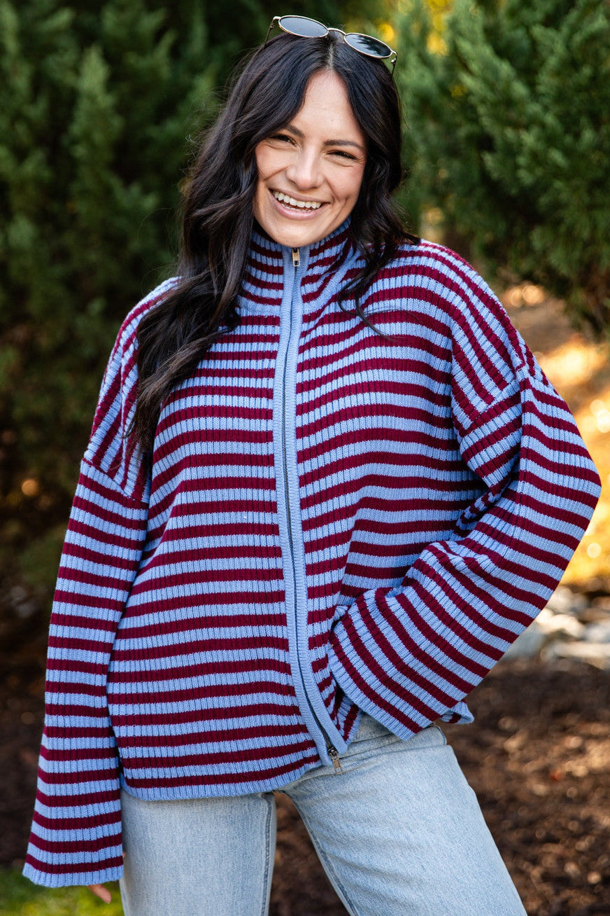 A woman smiling outdoors wearing a red and blue horizontally striped zip-up fleece jacket with a light blue zipper, paired with light-wash jeans.