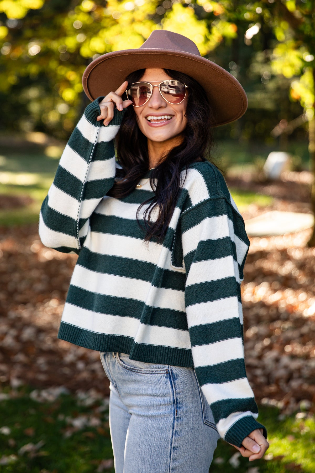 A woman wearing a dark green and white striped knit sweater, a brown wide-brim hat, and light-wash jeans, standing outdoors in a park with trees and fallen leaves in the background.
