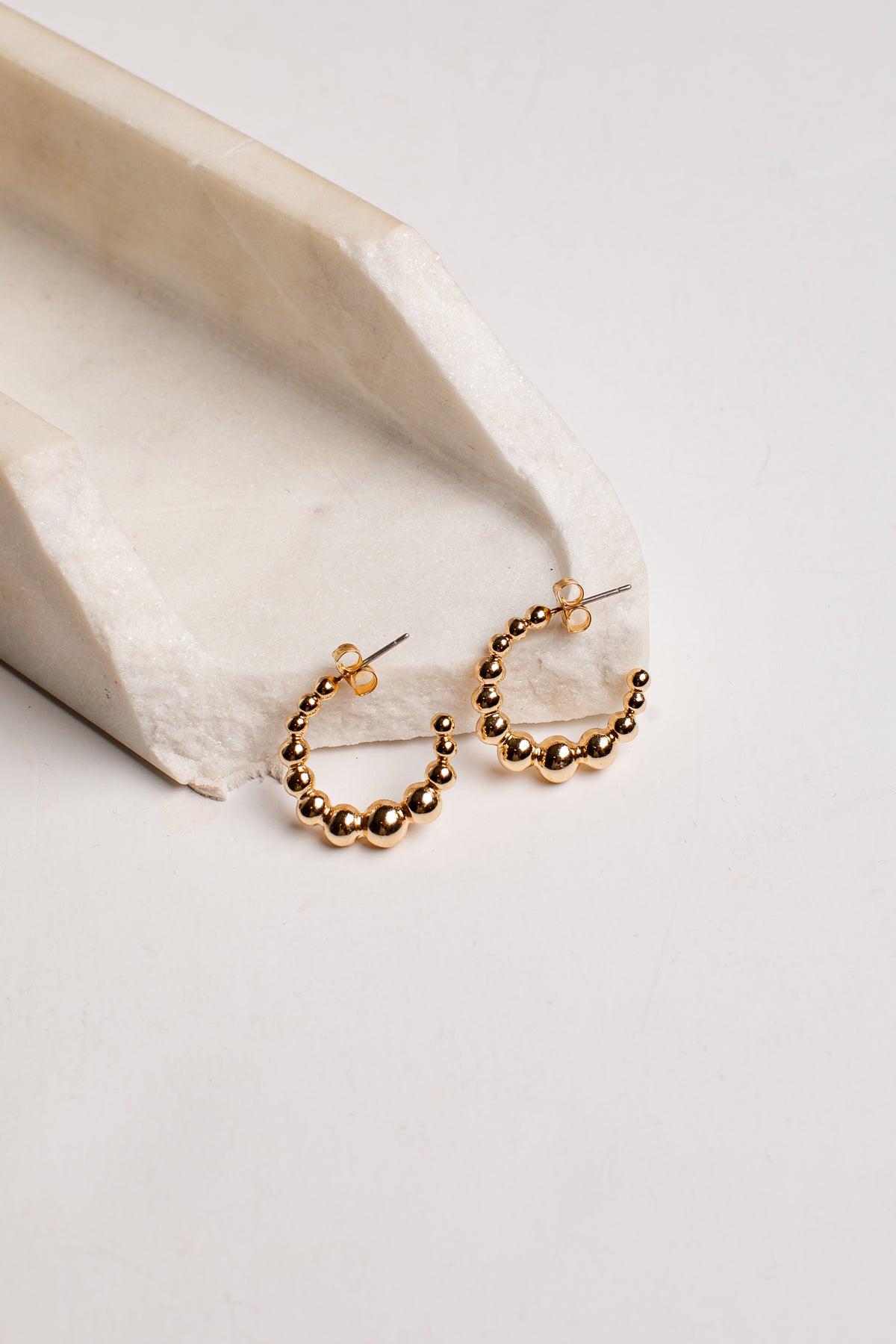 A pair of small gold hoop earrings with a beaded, textured design, resting on a white marble tray against a clean white background.
