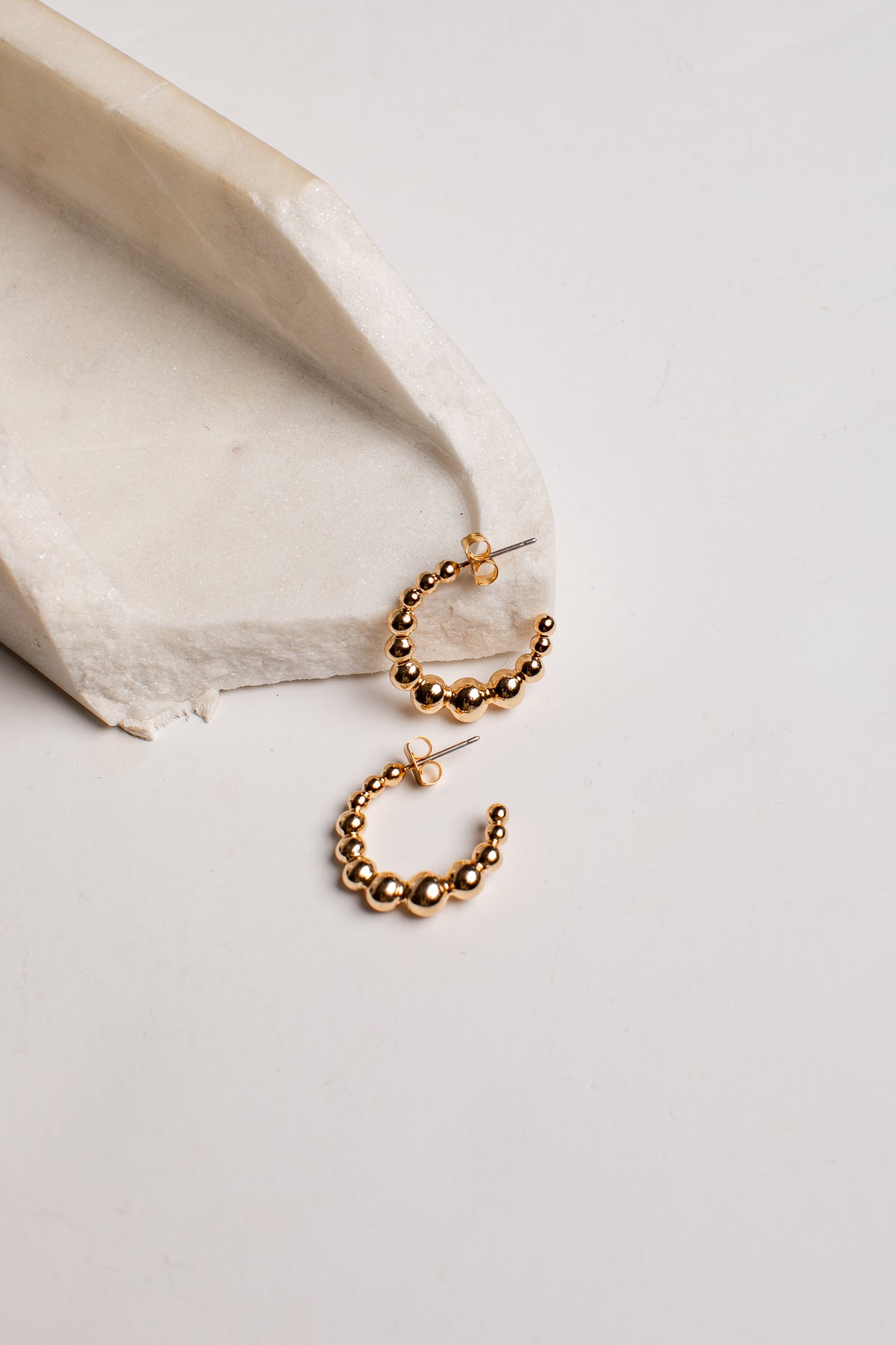 A pair of gold hoop earrings with a beaded, textured finish, displayed on a light gray surface next to a textured white stone prop.