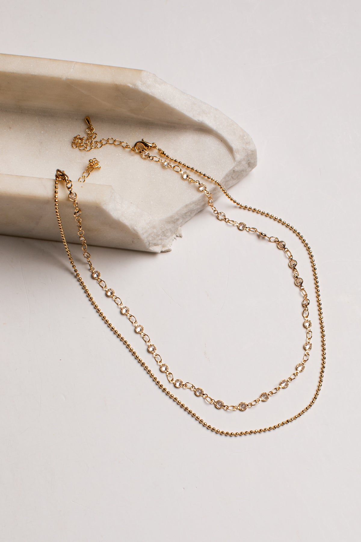 A delicate gold chain necklace draped over a textured white stone slab on a clean white background.
