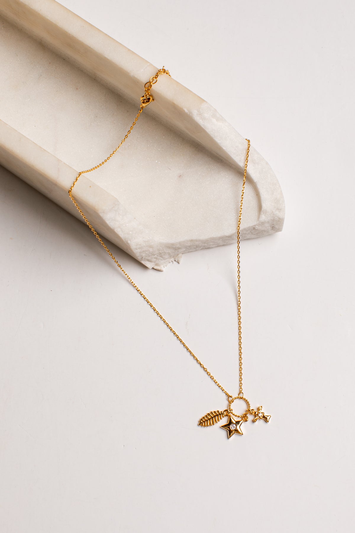 A delicate gold chain necklace with a small celestial pendant featuring a star, crescent moon, and feather, displayed on a light beige stone prop against a clean white background.