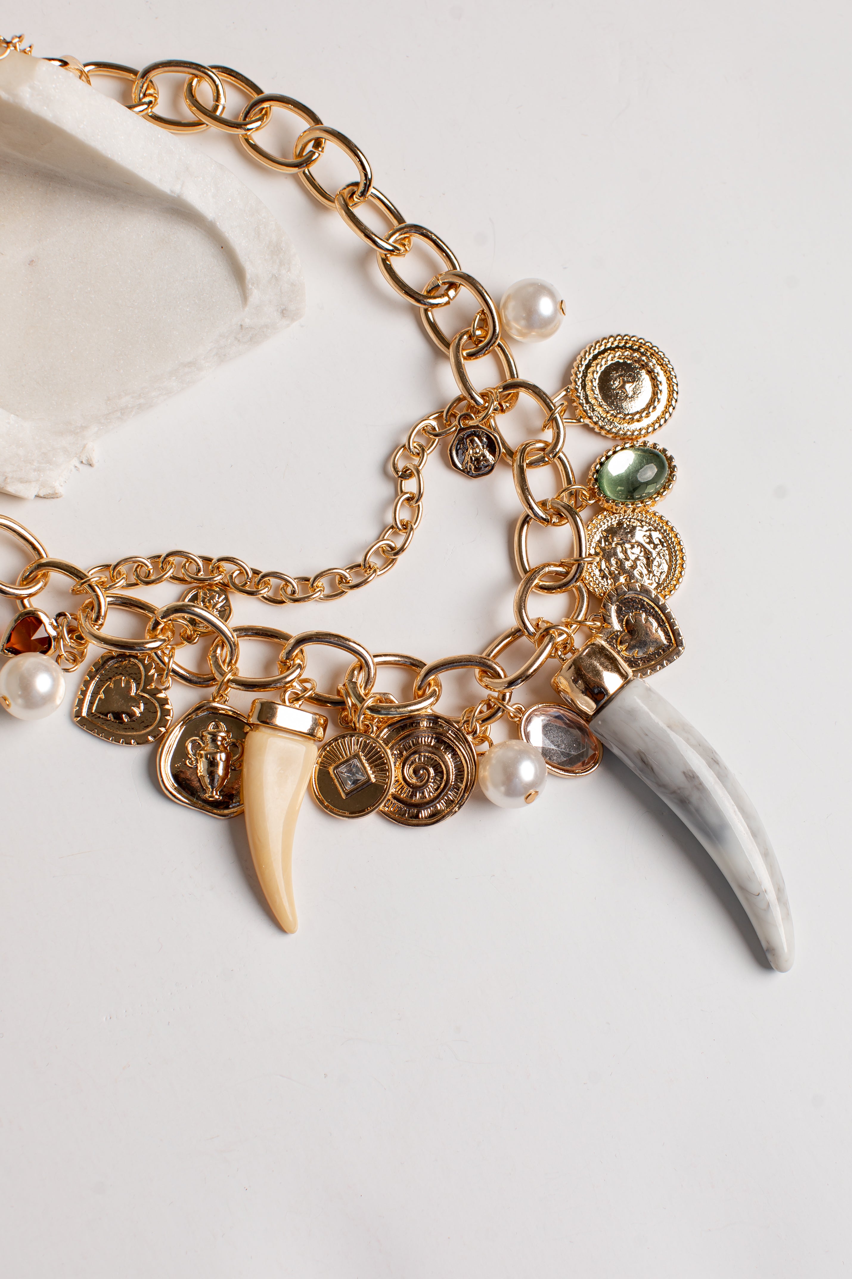 Gold-tone charm bracelet featuring a chunky chain, multiple coin charms, a cream-colored tooth, a white bone fragment, and small pearl beads on a light background.