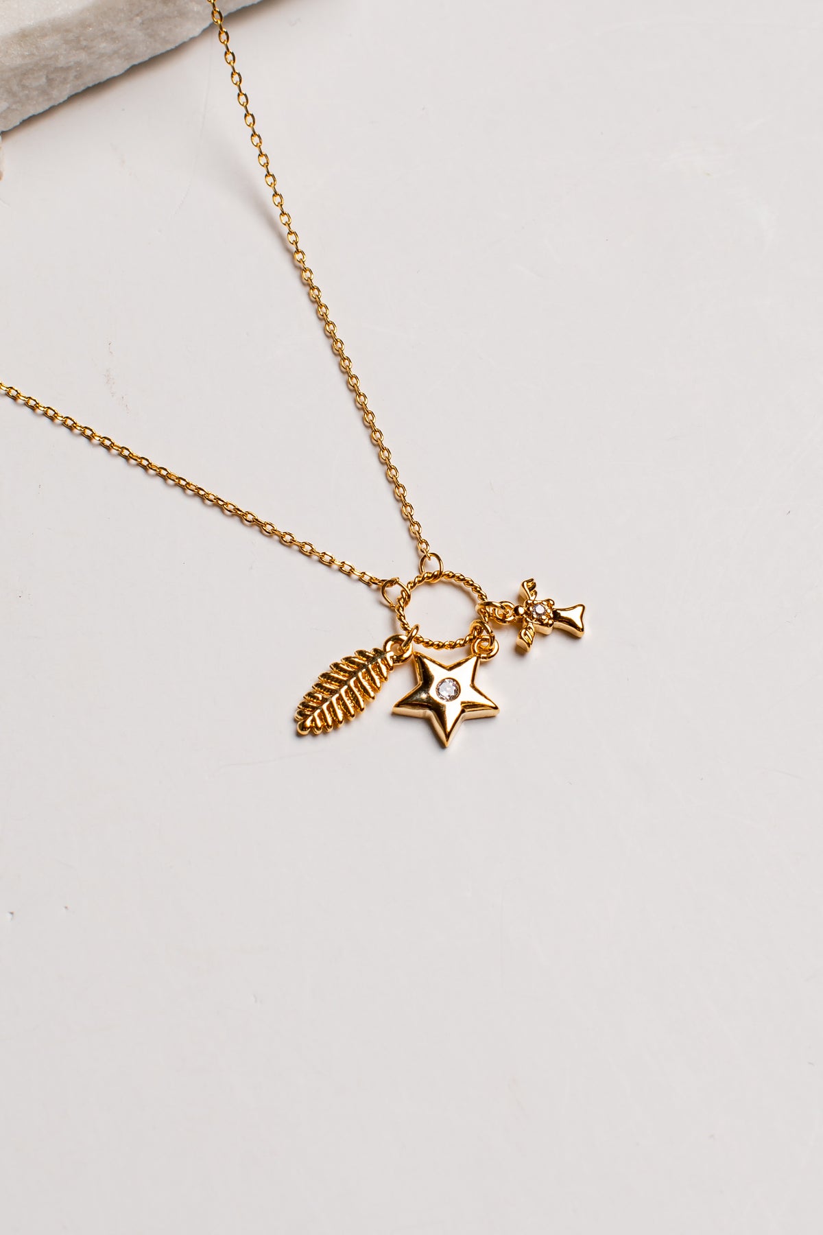 A delicate gold chain necklace featuring three charms: a textured feather, a star with a small clear gemstone, and a tiny bow and arrow, displayed on a light off-white background.