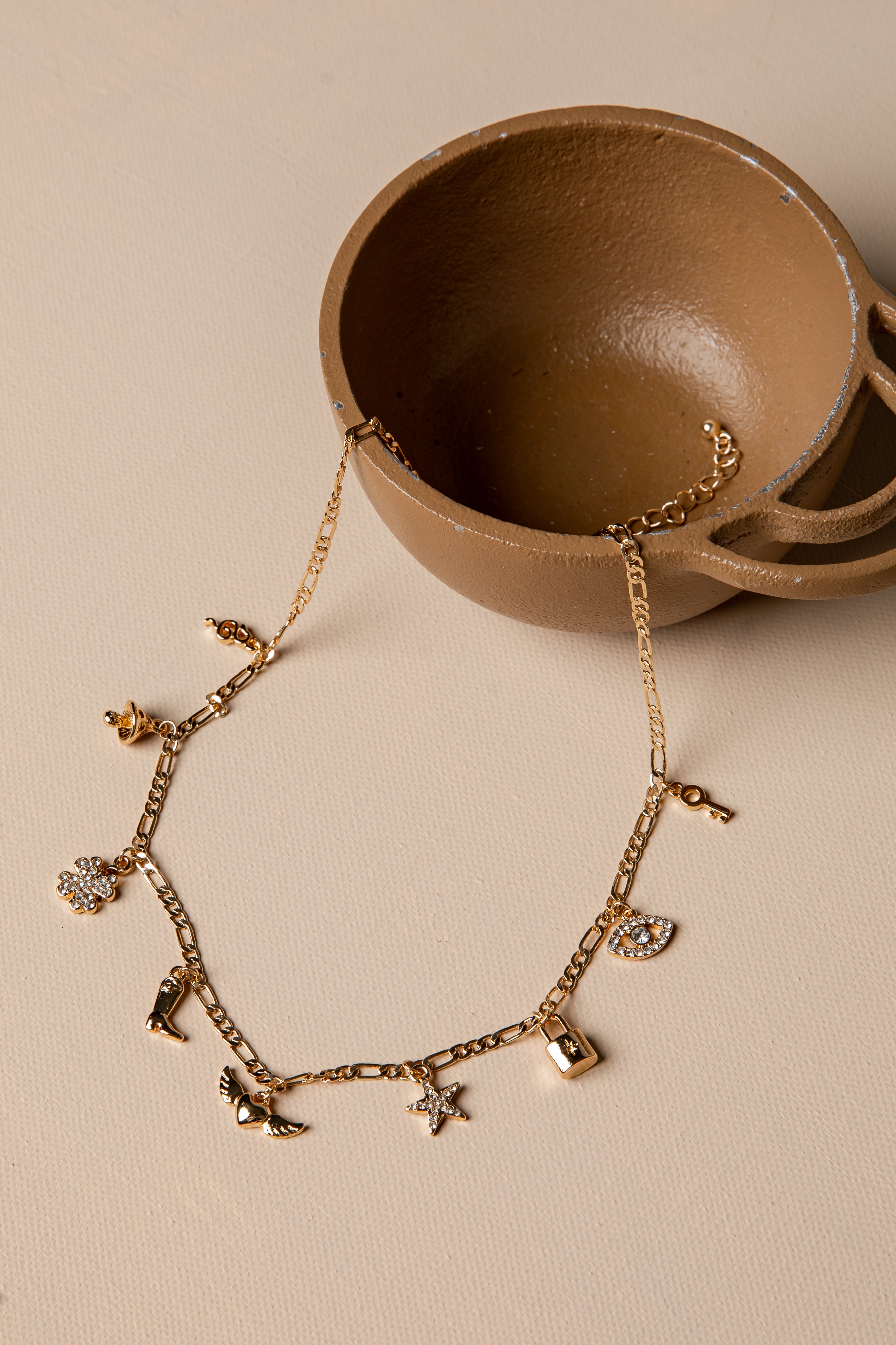Gold necklace with charms on a beige surface with a brown ceramic dish.