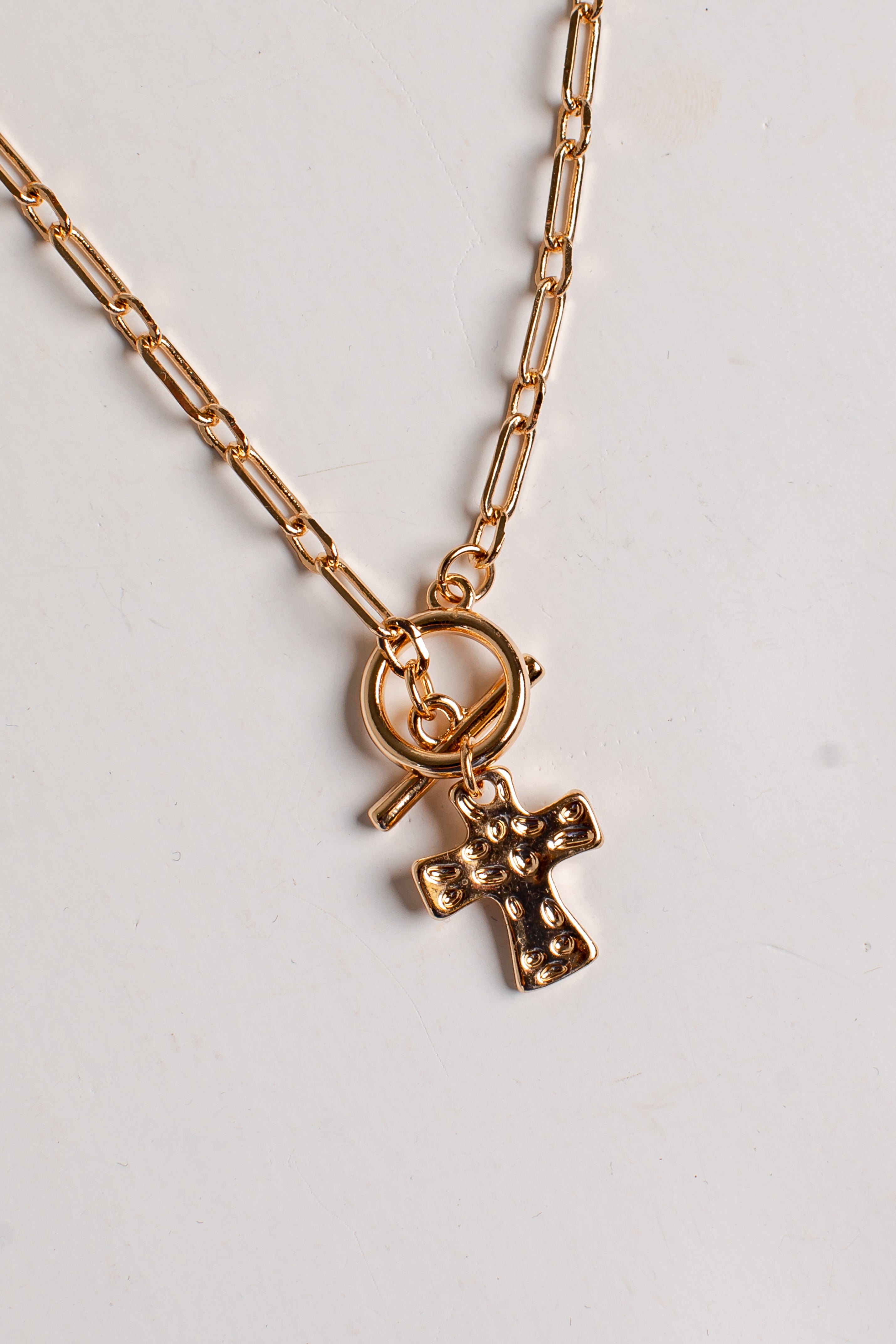 A gold-toned paperclip chain necklace with a small cross pendant featuring tiny clear rhinestones, displayed on a light off-white background.