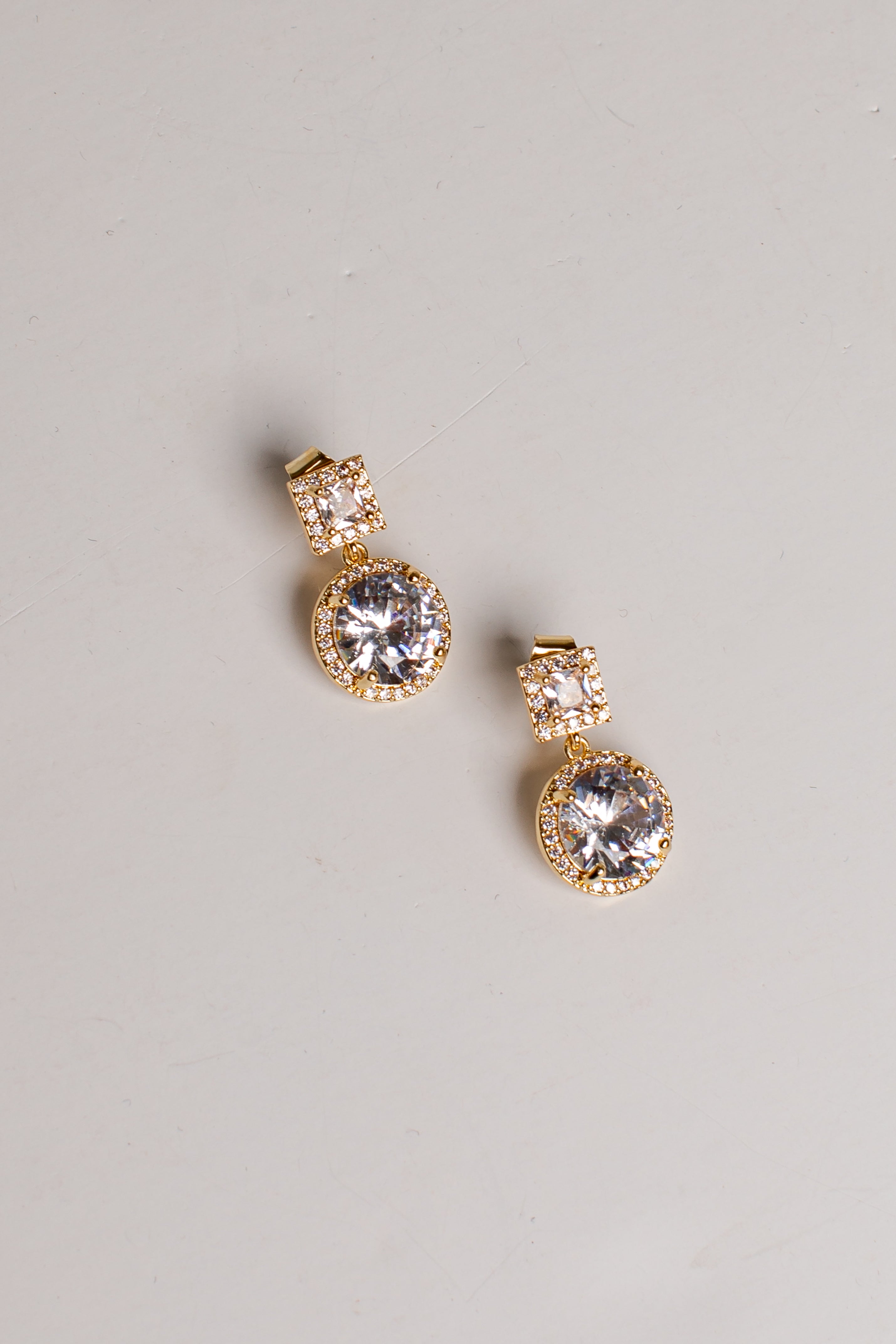 A pair of gold drop earrings with large clear round gemstones and smaller clear stones above them, photographed on a plain light gray background.