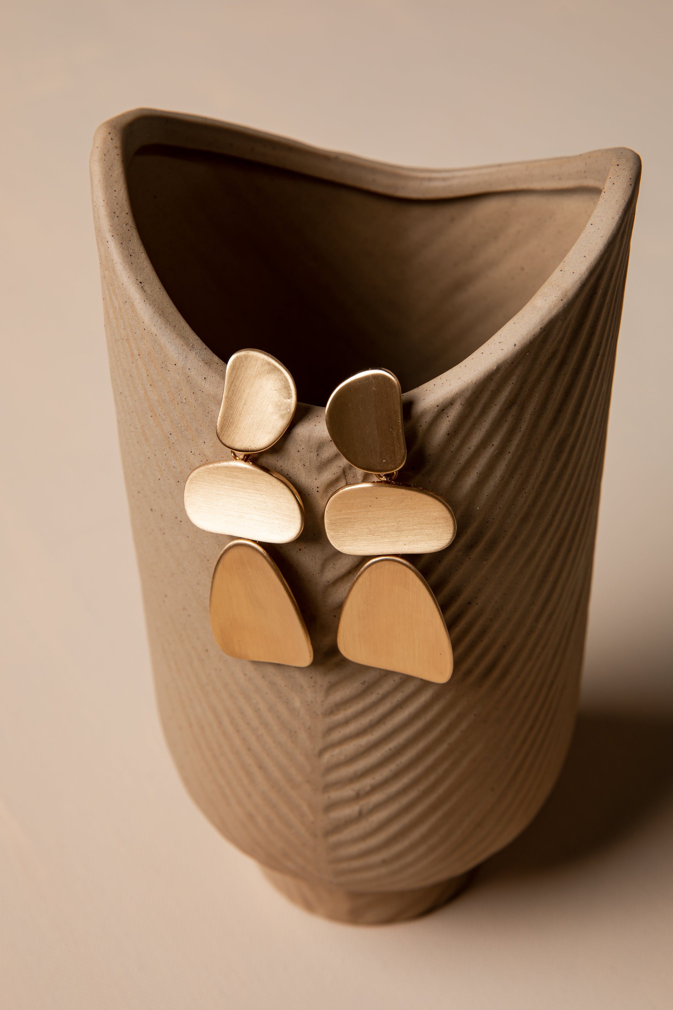 Gold earrings displayed on a textured beige vase against a matching background
