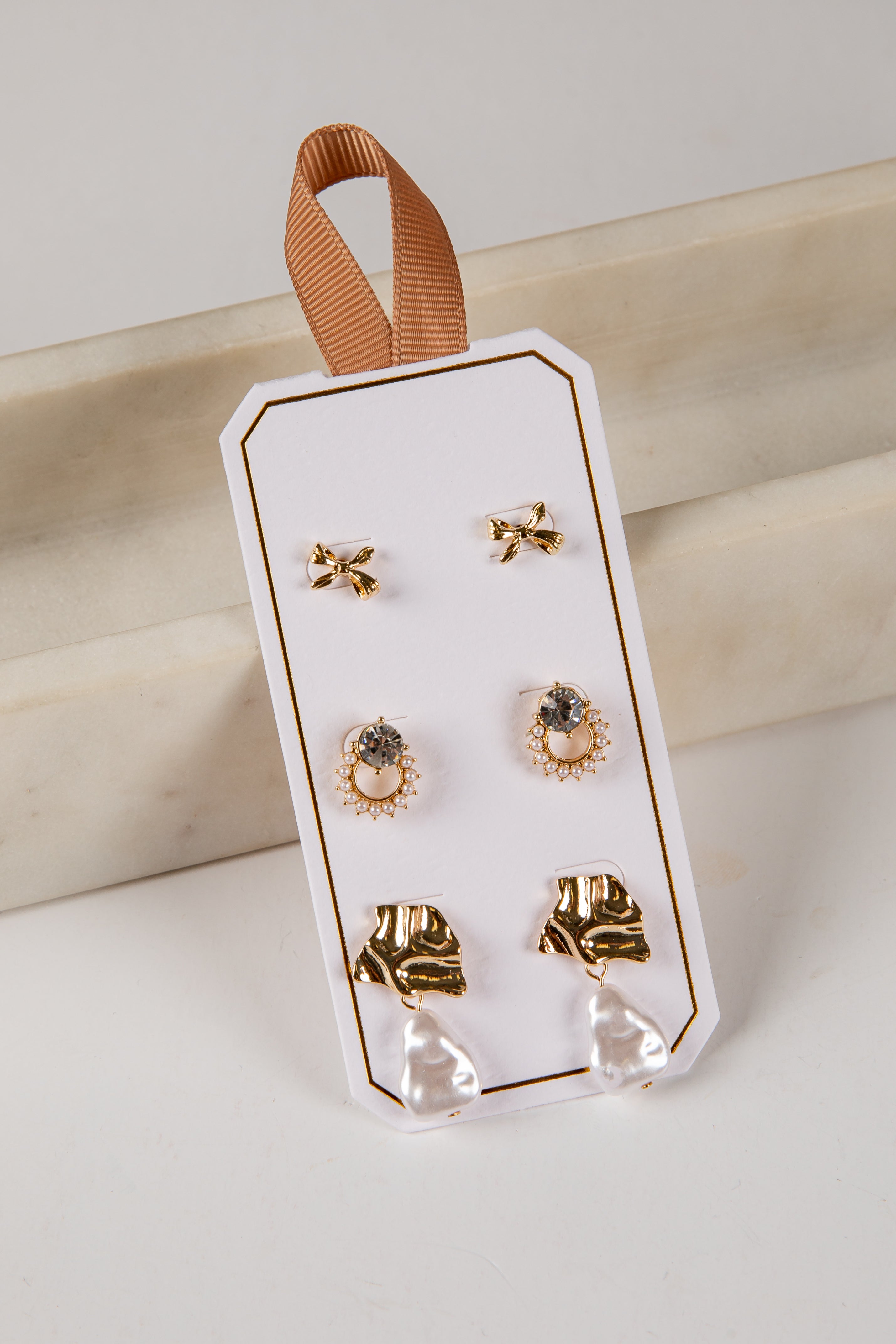 Set of gold earrings on a clear plastic card with a brown ribbon, against a light background.