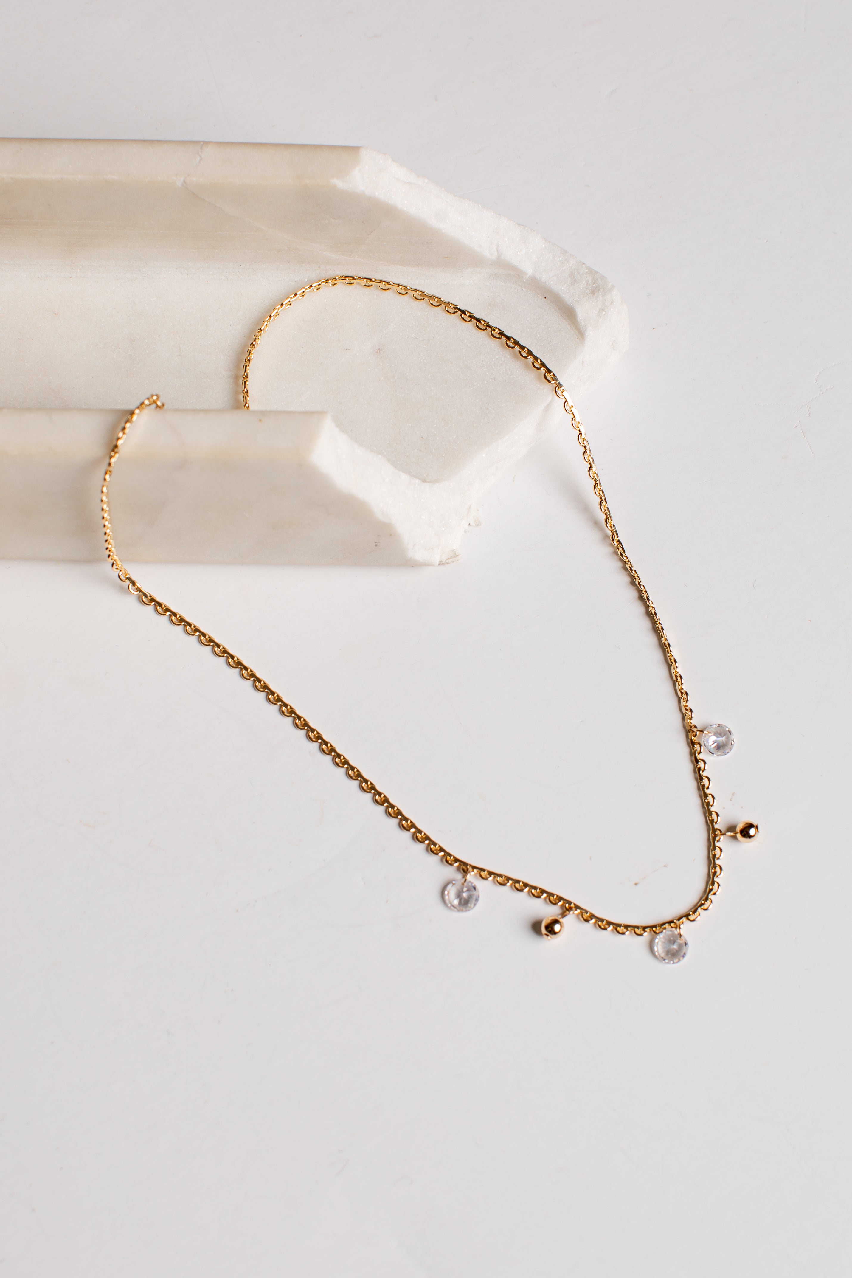 A delicate gold chain necklace with small clear crystal pendants and tiny gold beads, displayed on a white surface next to a cream-colored stone prop.