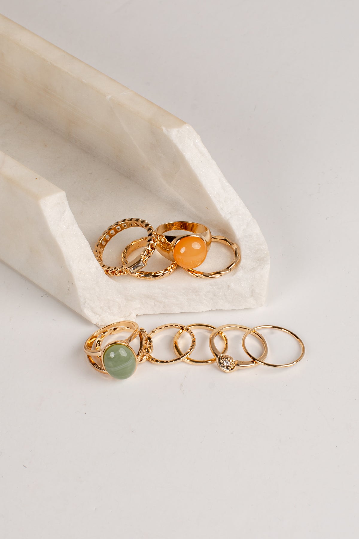 A set of gold-toned rings displayed on a white marble stand and flat surface, featuring textured bands, a smooth orange stone, a green stone, and a small clear gemstone.