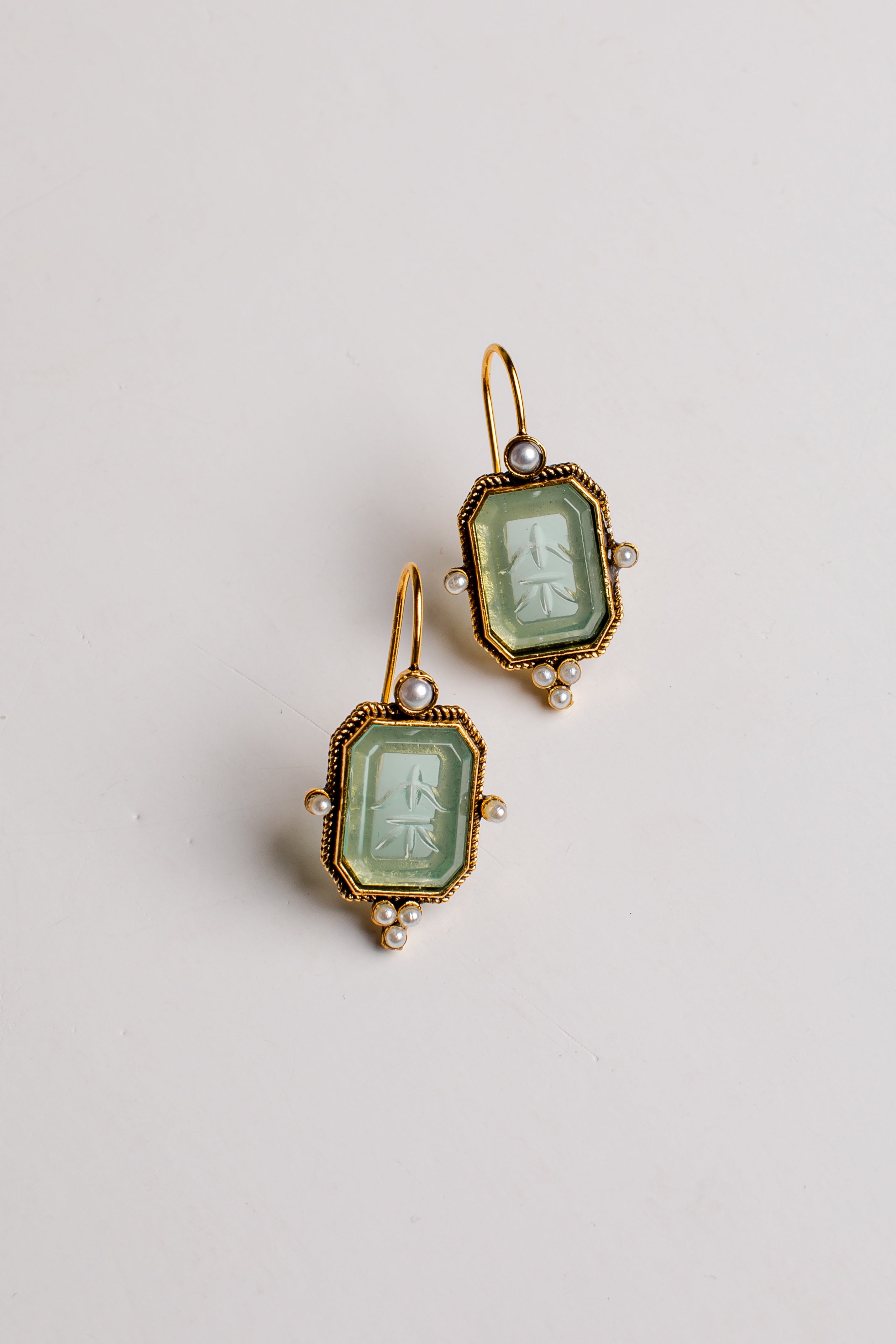 A pair of antique-style gold drop earrings featuring light green carved glass panels with classical figures, accented with small white pearls, shown on a plain light background.
