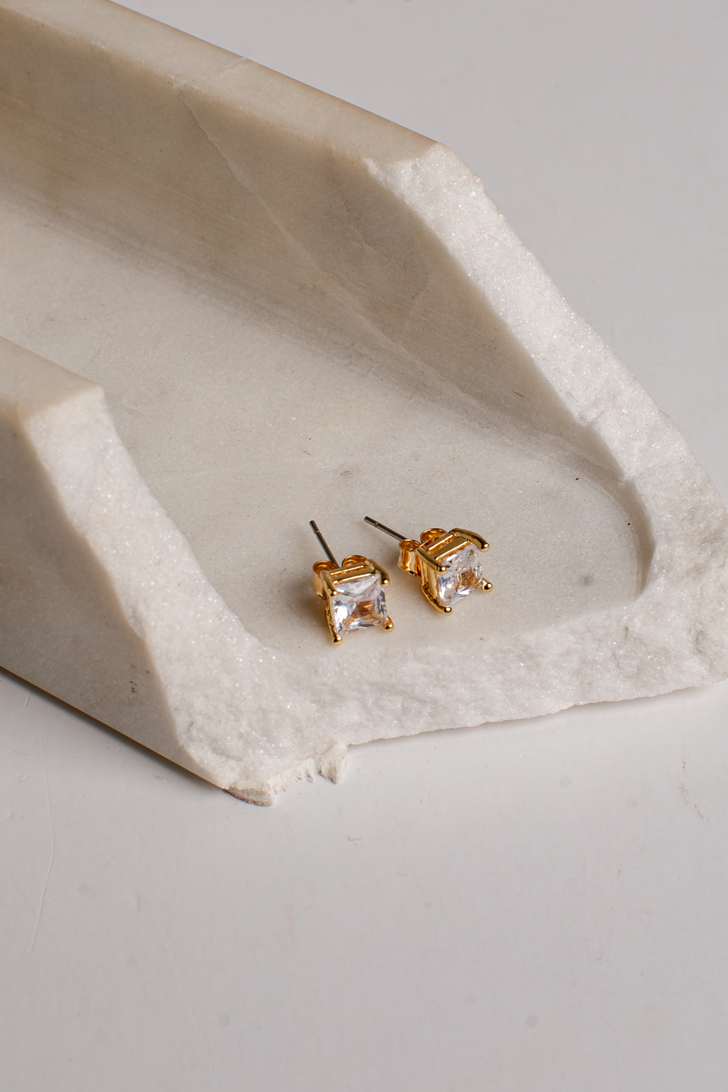 A pair of gold stud earrings with clear gemstones, resting on a textured, off-white stone tray against a light gray background.