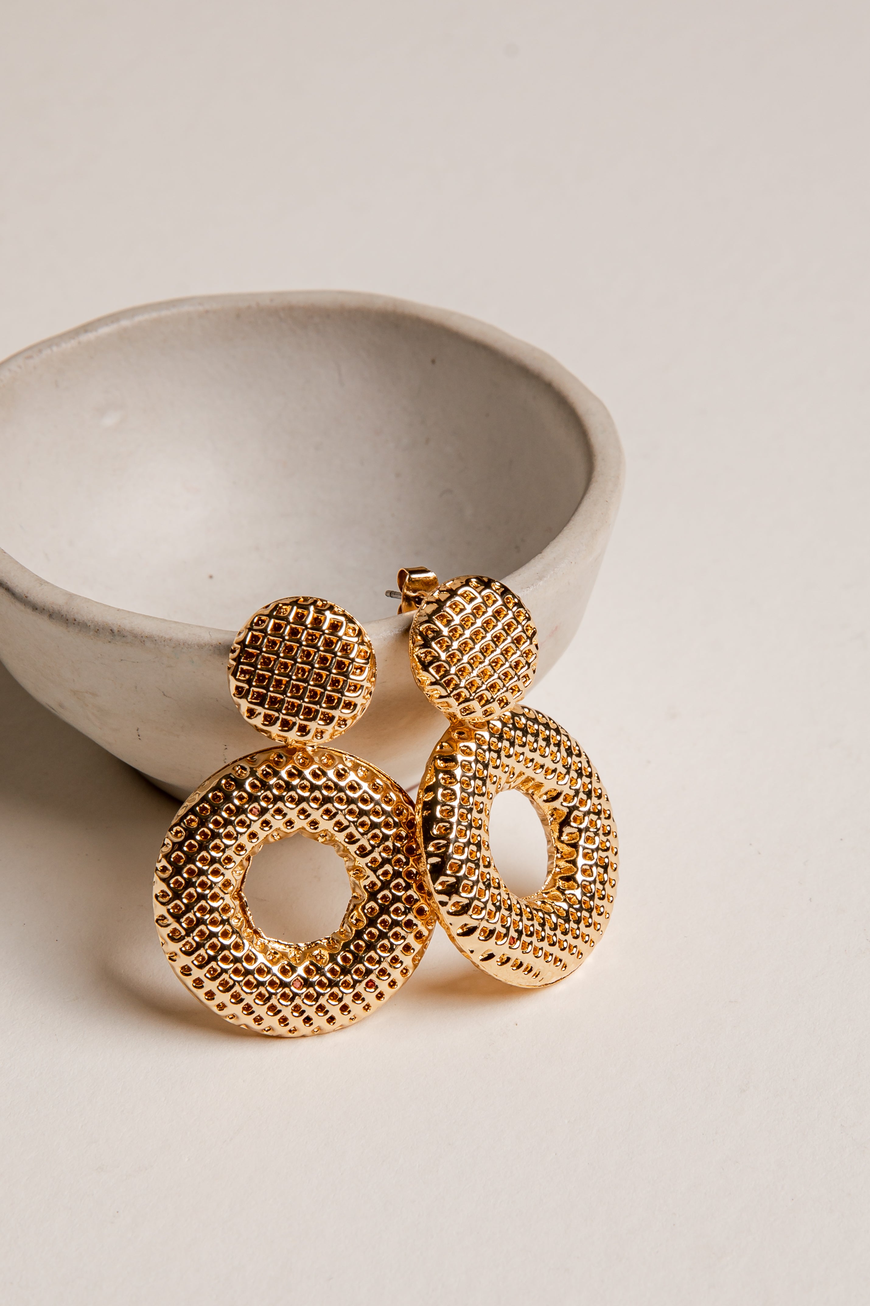 Gold earrings on a light beige surface with a ceramic bowl in the background