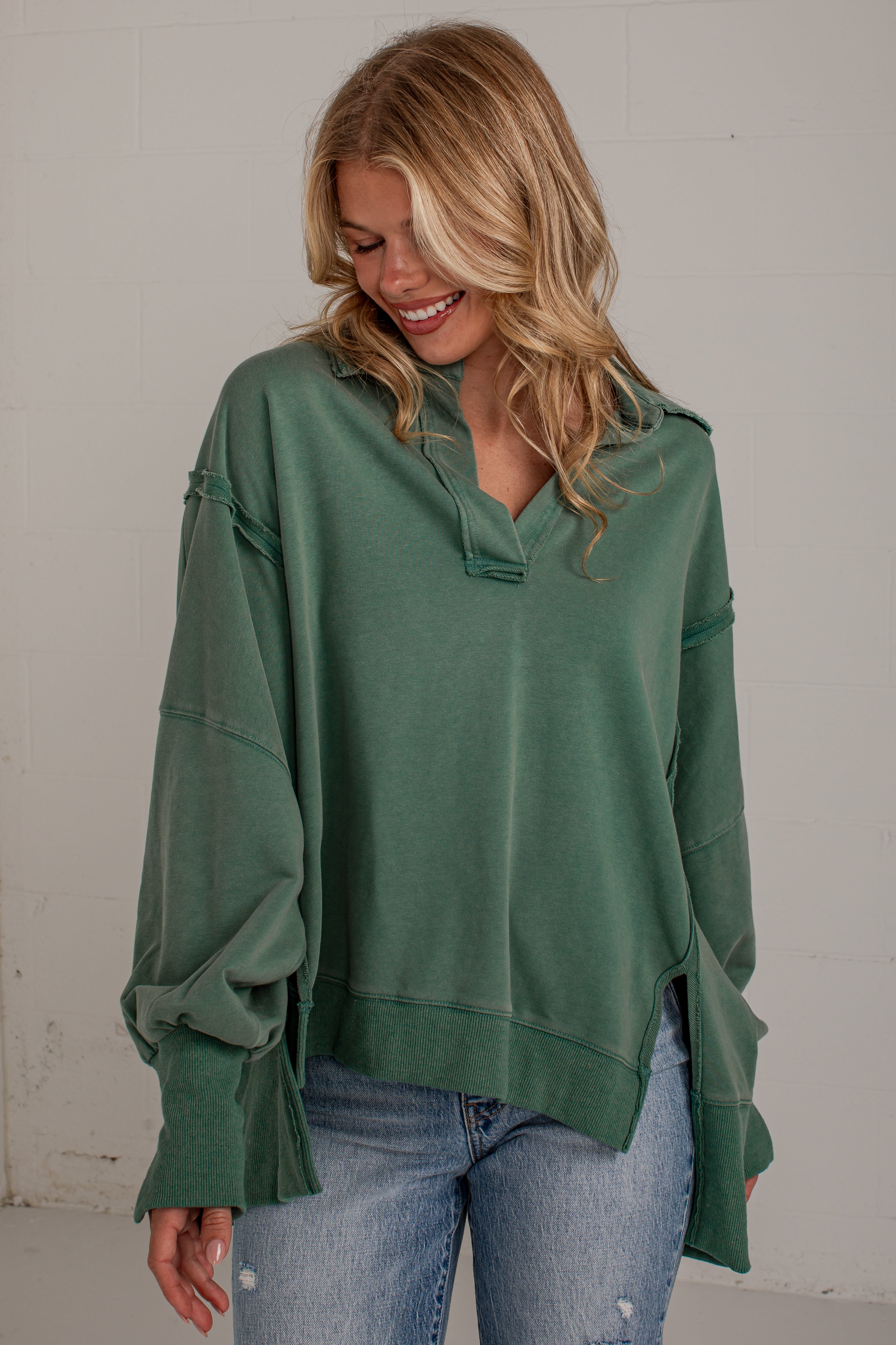 A woman wearing an oversized sage green knit top with unique ruffled cuffs and a split neckline, paired with light-wash blue jeans, standing against a light gray wall.