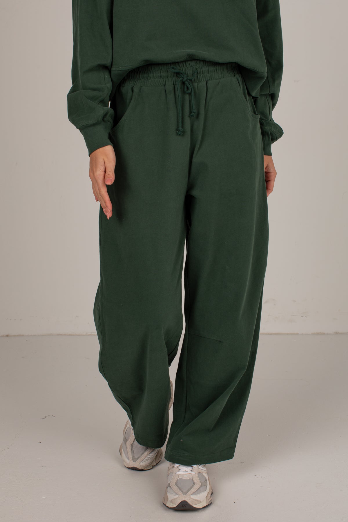 A person wearing dark green wide-leg sweatpants with an elastic drawstring waist, paired with a matching dark green sweatshirt and white sneakers, standing against a plain light gray studio background.